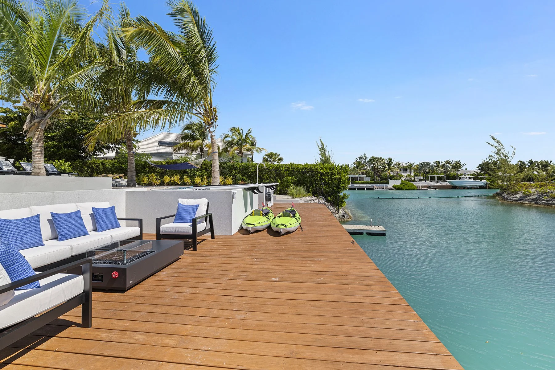A waterfront wooden deck with white outdoor furniture and blue pillows, two green kayaks, palm trees, and a marina with boats on a sunny day.