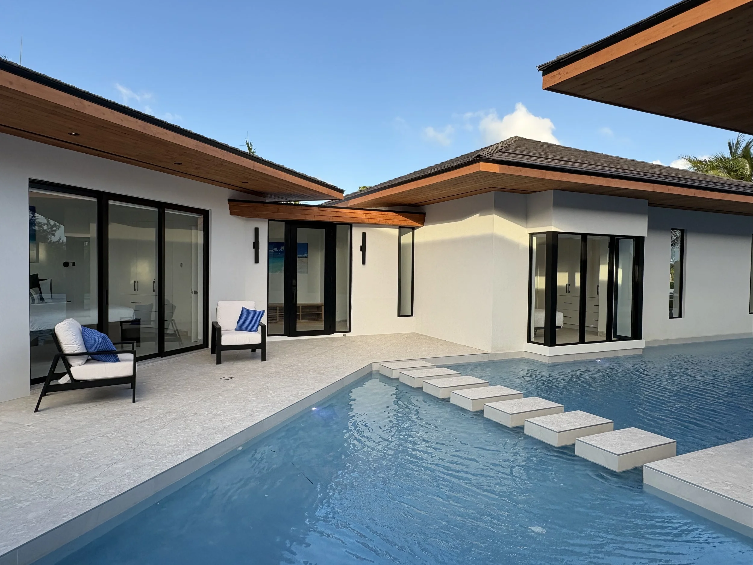Modern house with large glass sliding doors, a swimming pool with stepping stones, and outdoor seating with white cushions and blue pillows. Clear sky with some clouds in the background.