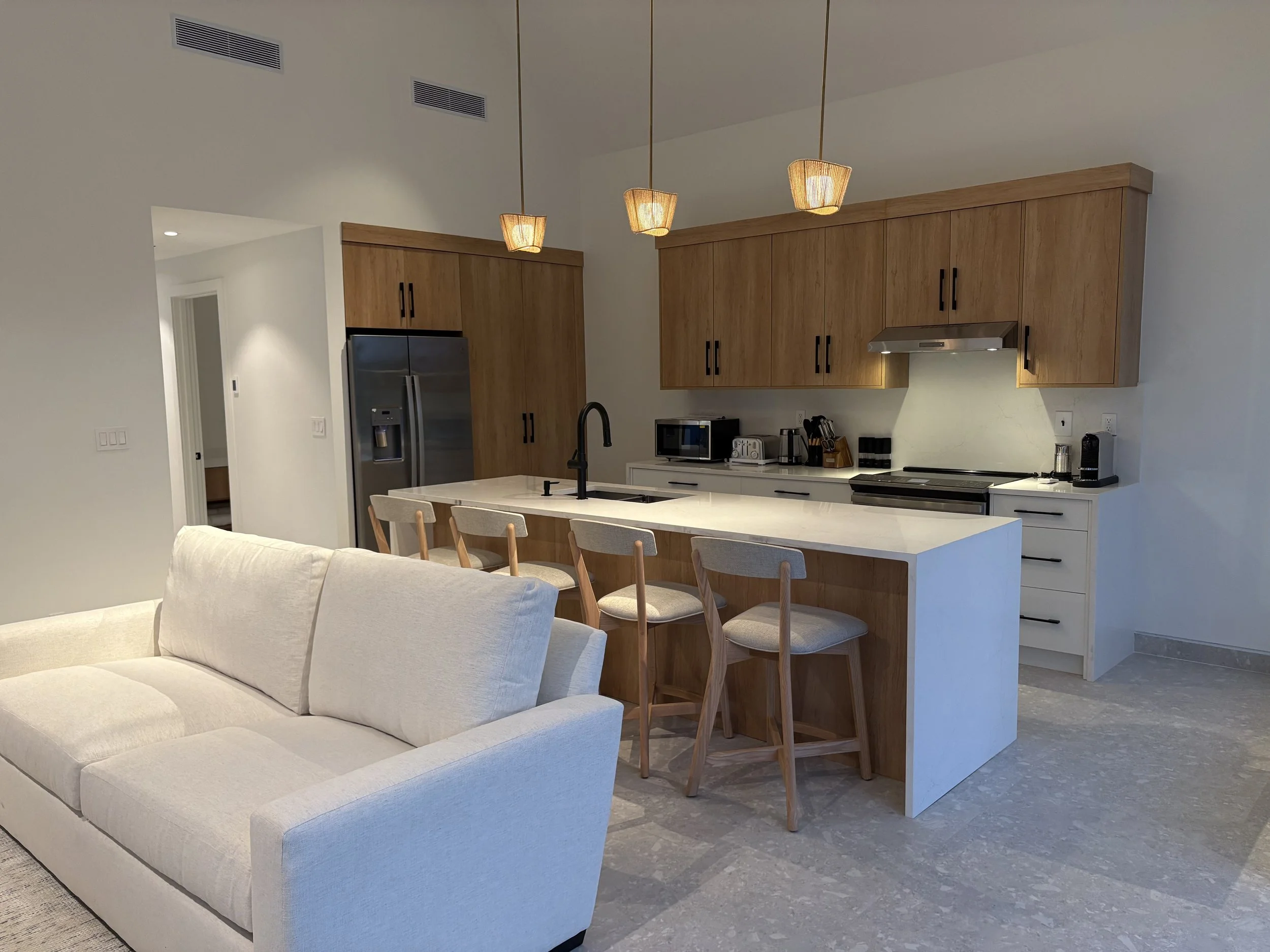 Modern open-concept kitchen with a white island, wooden cabinets, stainless steel appliances, and three pendant lights, adjacent to a white sofa in a living area.