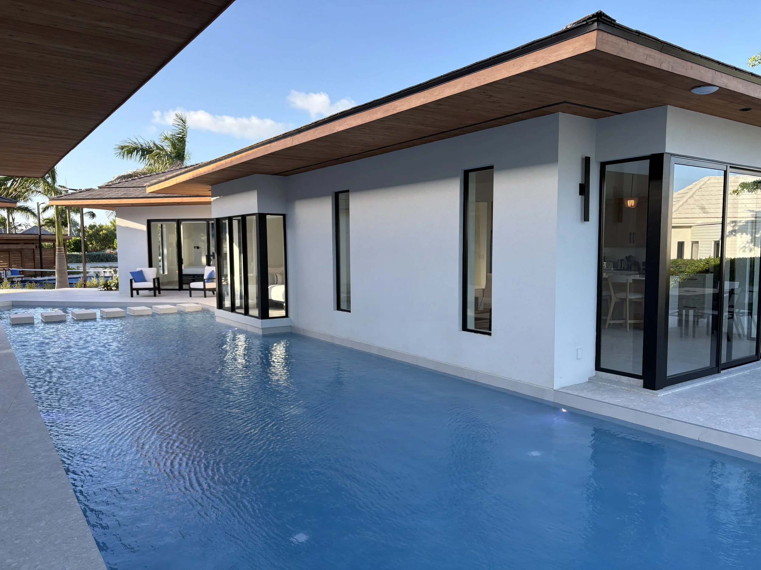 Modern house with a swimming pool and outdoor seating area, featuring large glass doors and windows, wooden overhangs, and tropical palm trees in the background.