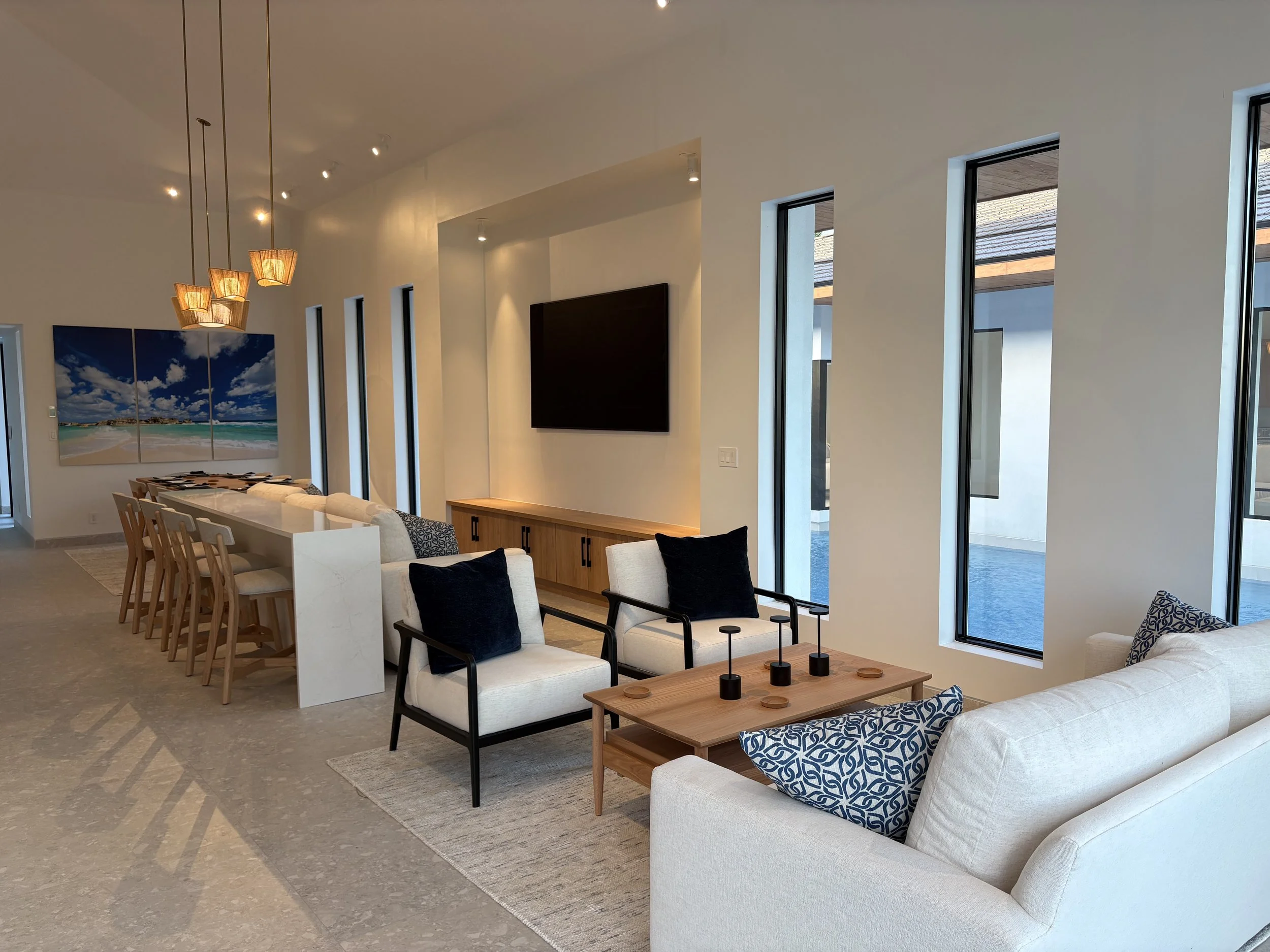 Modern living room with beige furniture, black and blue pillows, wooden coffee table, TV wall, and large windows with ocean view.