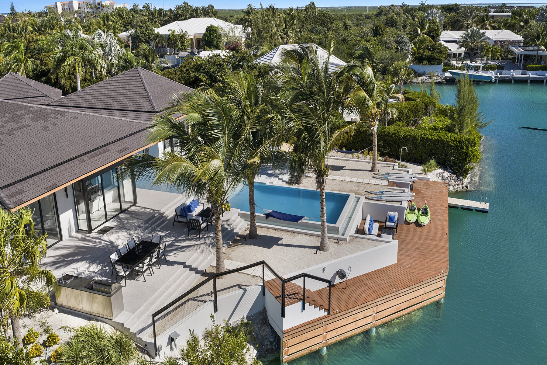Aerial view of a modern waterfront house with a swimming pool, palm trees, outdoor seating, and a dock with kayaks on clear blue water.