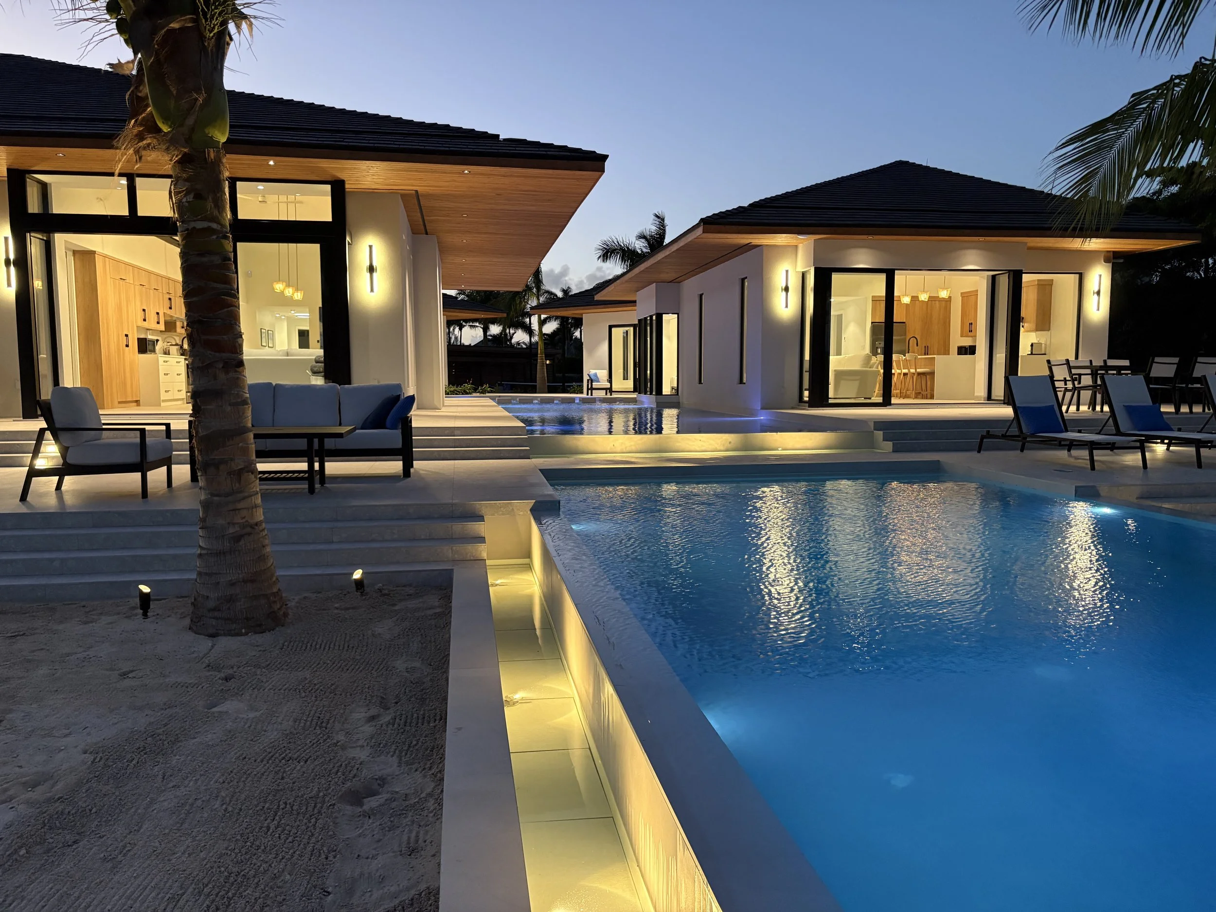 Luxury modern house with illuminated pool, outdoor seating, and large glass doors showing interior kitchens at dusk.