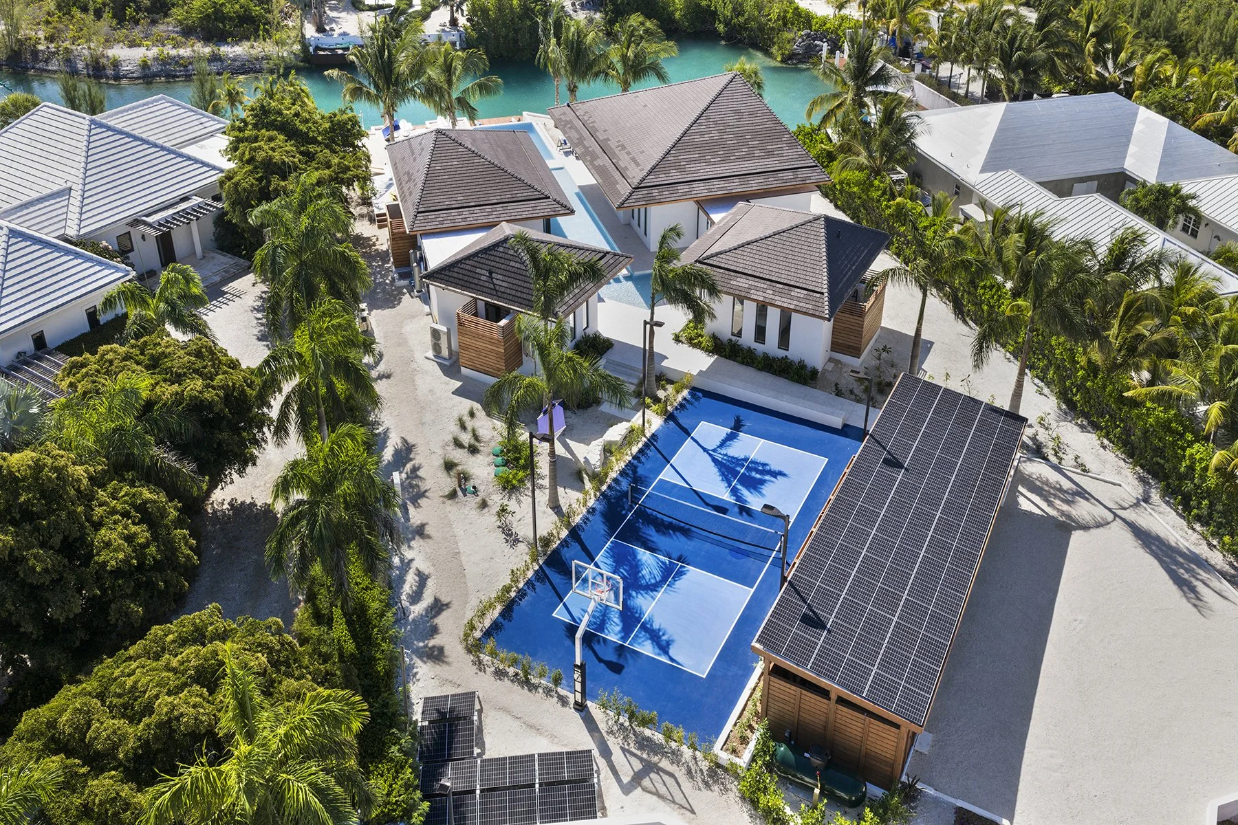 Aerial view of a tropical resort featuring a swimming pool, tennis court, beach area, and surrounding lush greenery and palm trees.
