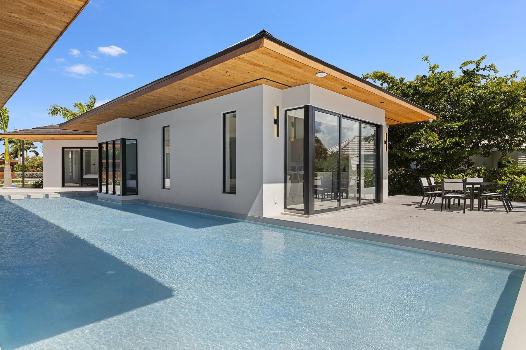 Modern house with a pool, outdoor dining area, and large windows, on a sunny day with a blue sky and trees in the background.