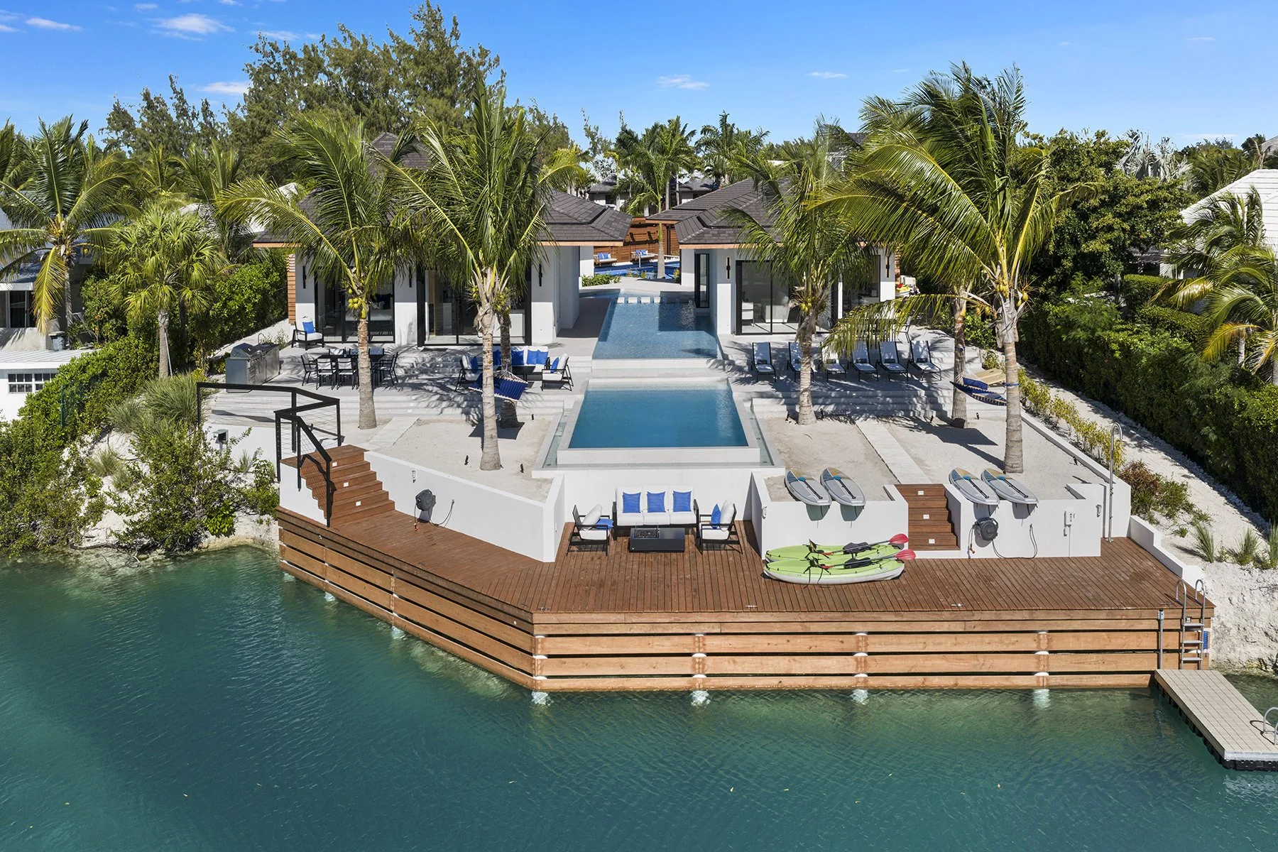 Aerial view of a modern beachfront villa with a swimming pool, lounge chairs, palm trees, and outdoor seating on a wooden deck overlooking the water.