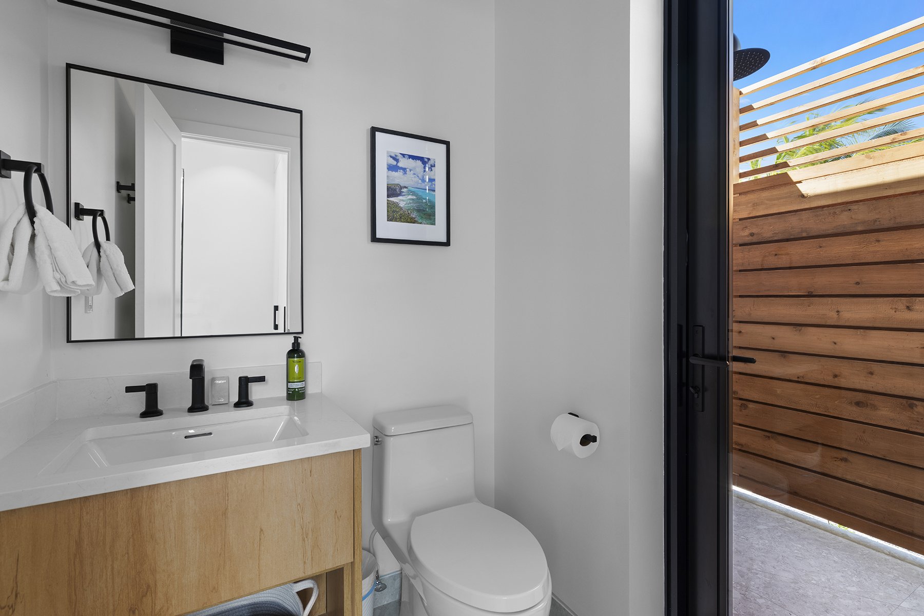 Modern bathroom with white walls, a wooden vanity with a white sink, black fixtures, and a large mirror. There is a framed picture on the wall, a toilet paper roll, and a bottle of hand soap. The door opens to an outdoor area with a wooden privacy fe