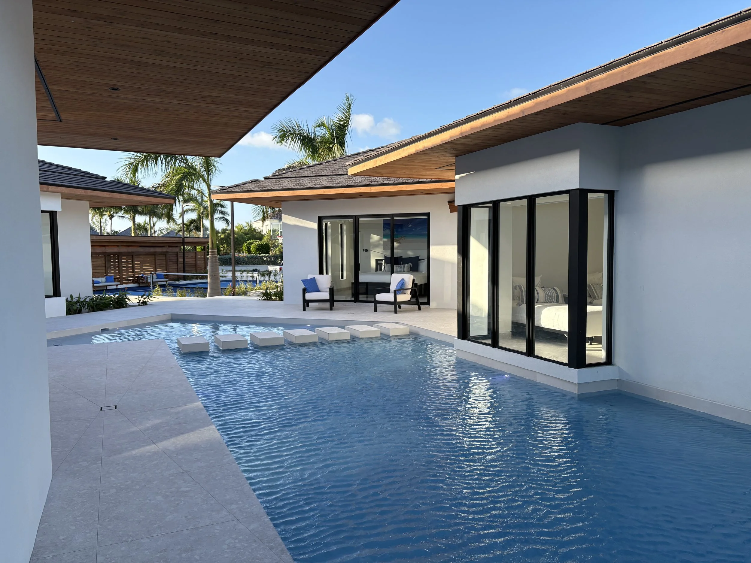 Modern house with a swimming pool, glass doors, outdoor chairs, and palm trees in a sunny backyard.