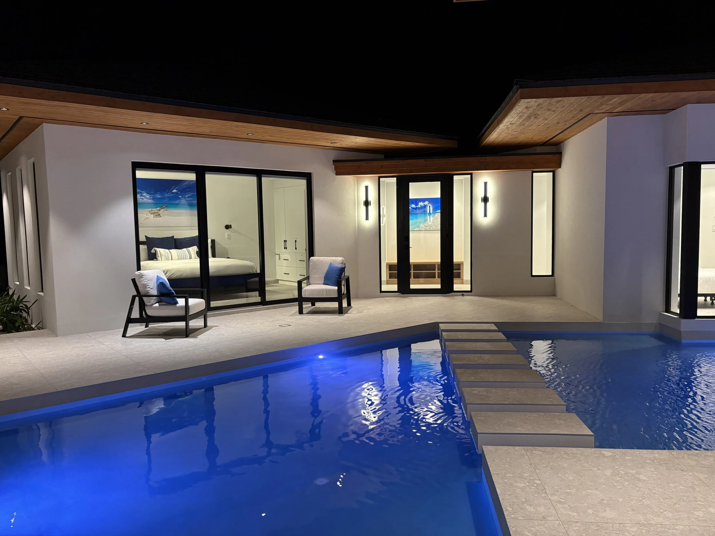 Night view of a modern house with a swimming pool, two chairs with cushions outside, and a bedroom with a large bed visible through glass doors.