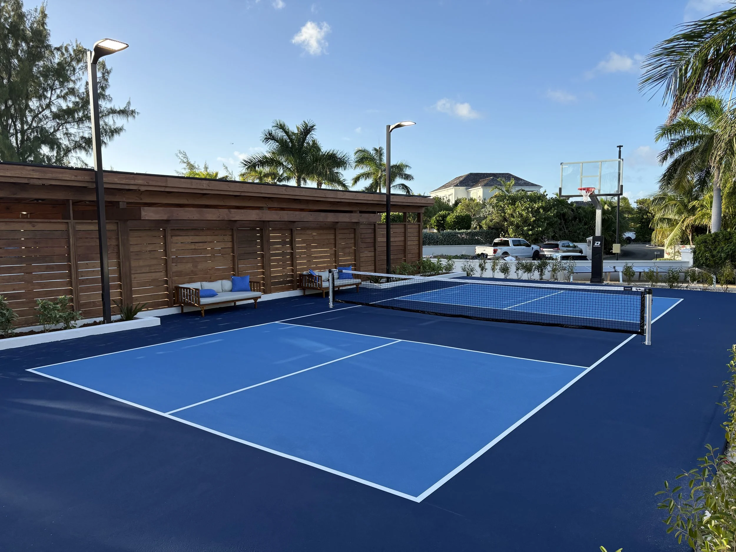 An outdoor blue tennis court with a net in the center, surrounded by green palm trees and benches with blue cushions, under a partly cloudy sky.