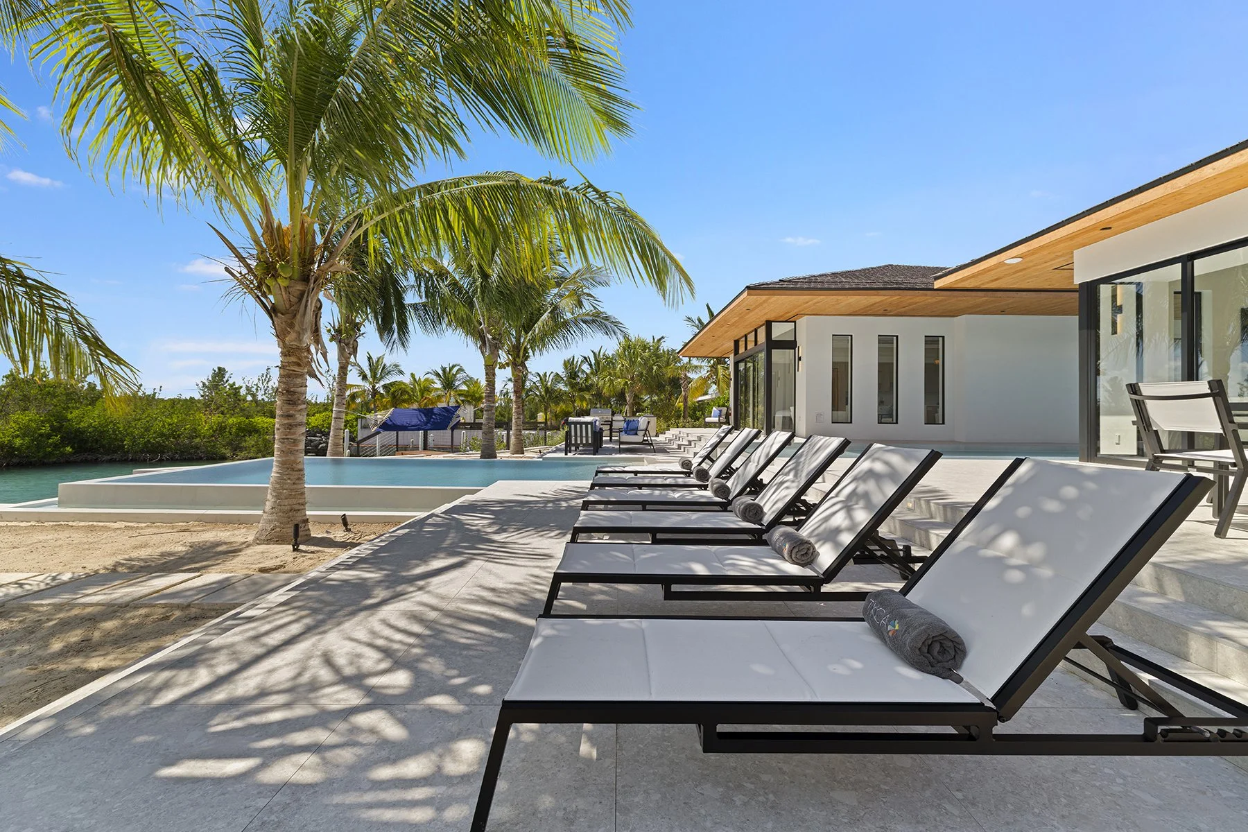Luxury modern house with a swimming pool, lounge chairs, palm trees, and outdoor furniture under a clear blue sky.