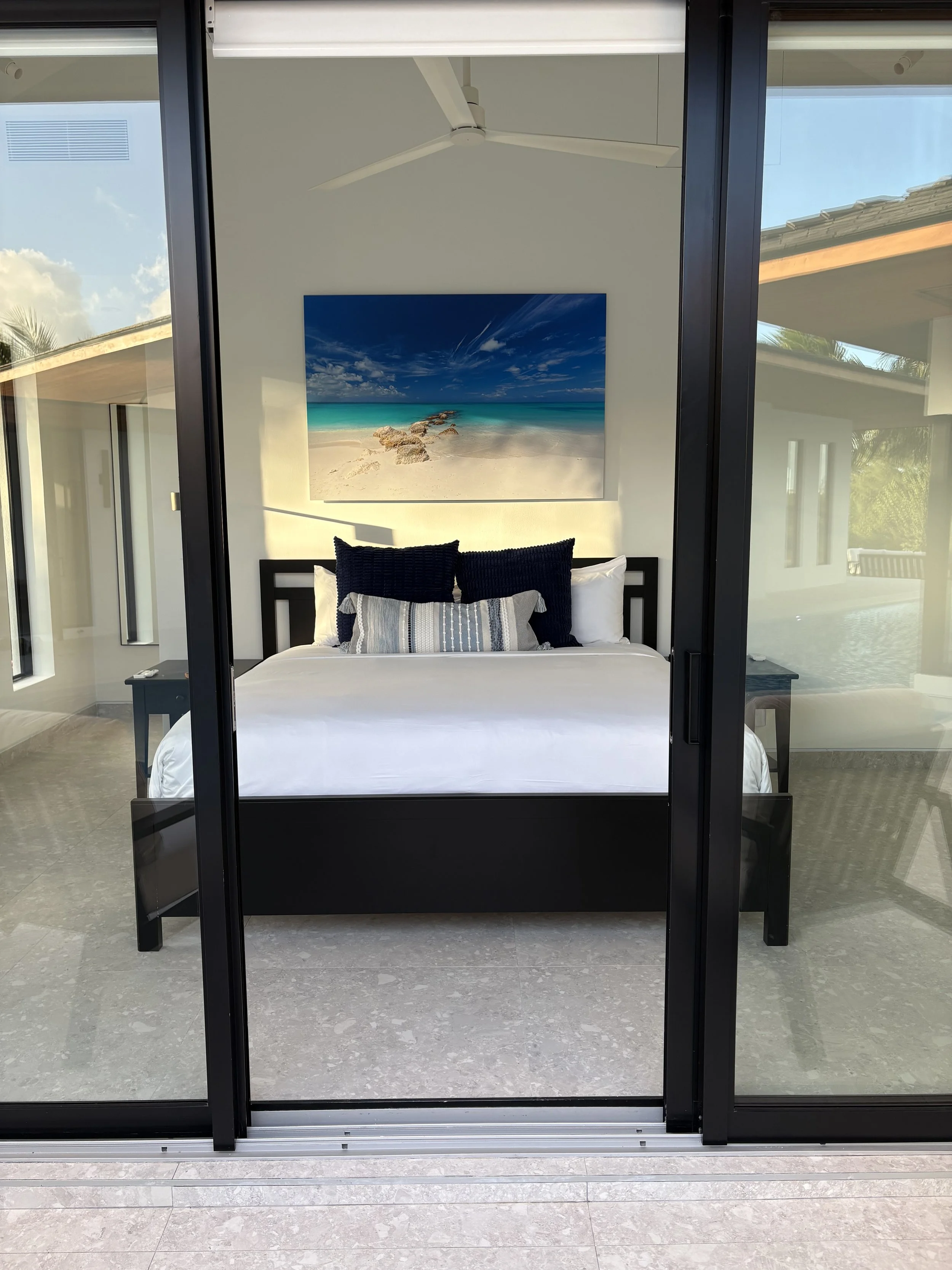View of a bedroom through glass sliding doors with a black bed frame, white bedding, and decorative pillows. A beach scene painting hangs above the bed, and a ceiling fan is visible.