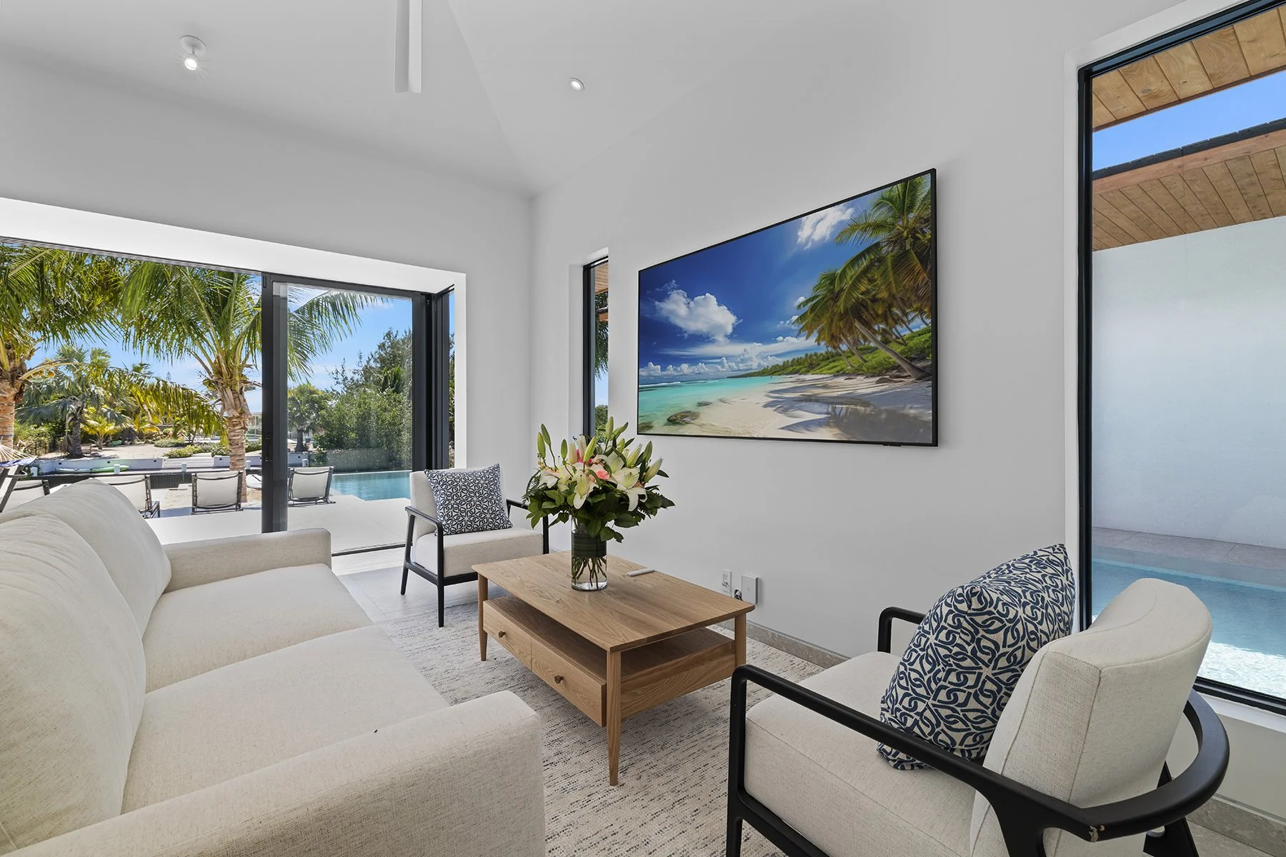 Modern living room with white walls, large glass sliding doors, and a view of a backyard with palm trees and pool. Furniture includes a beige sofa, two armchairs with patterned cushions, a wooden coffee table with a flower vase, and a large wall-moun