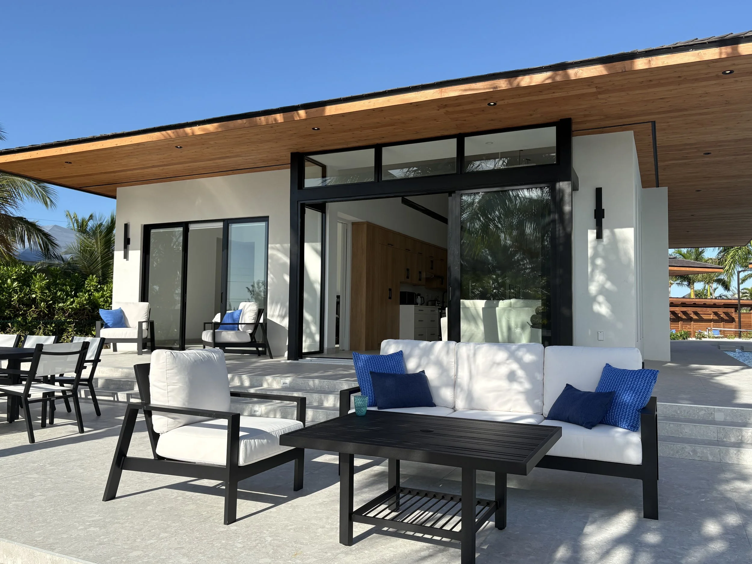 Modern outdoor patio with white cushioned seating, black wooden furniture, and a house with large glass sliding doors and windows, surrounded by tropical trees and clear blue sky.