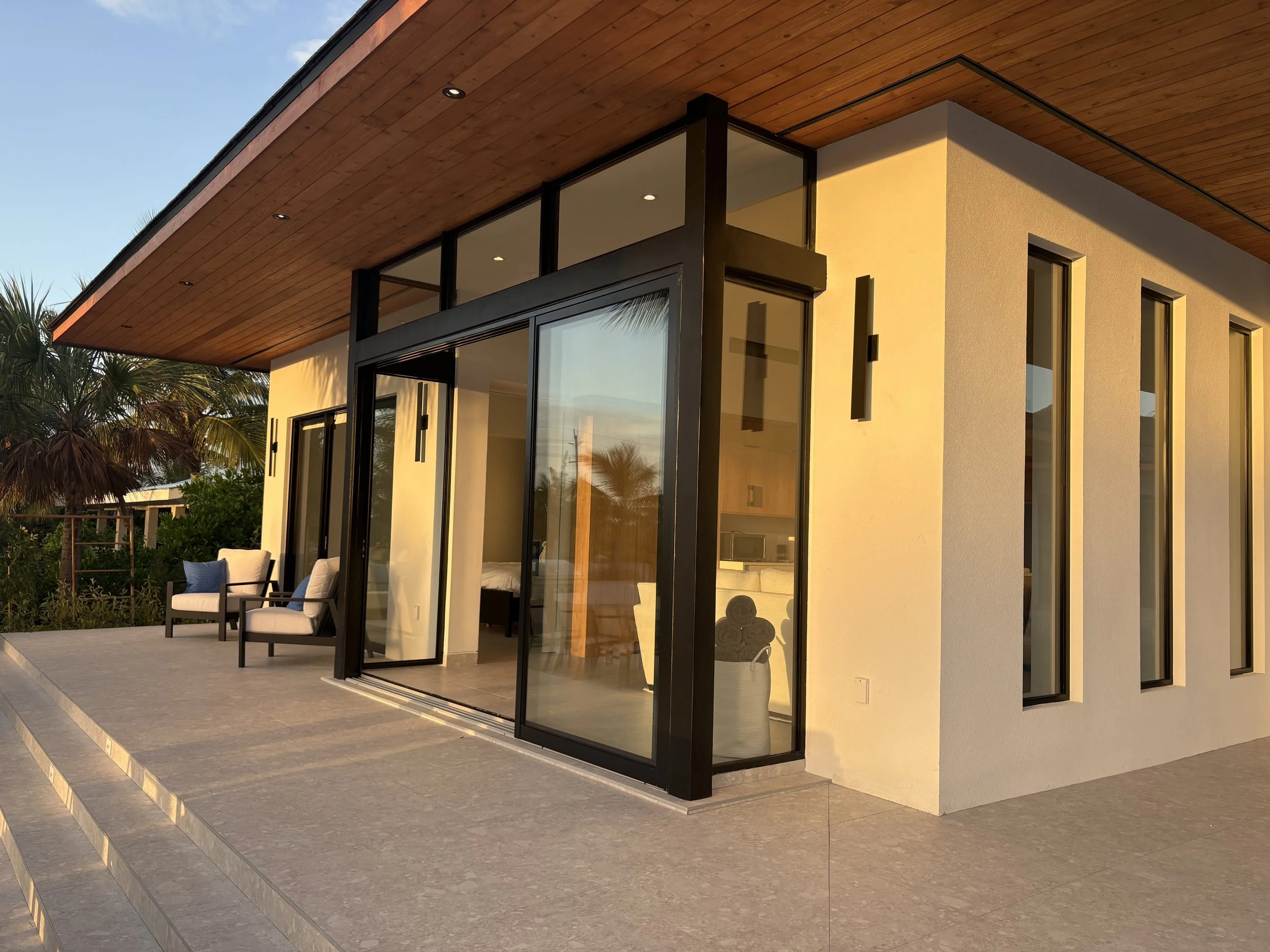 Modern house with a porch, large glass sliding doors, and multiple narrow windows, surrounded by greenery and palm trees, during sunset.