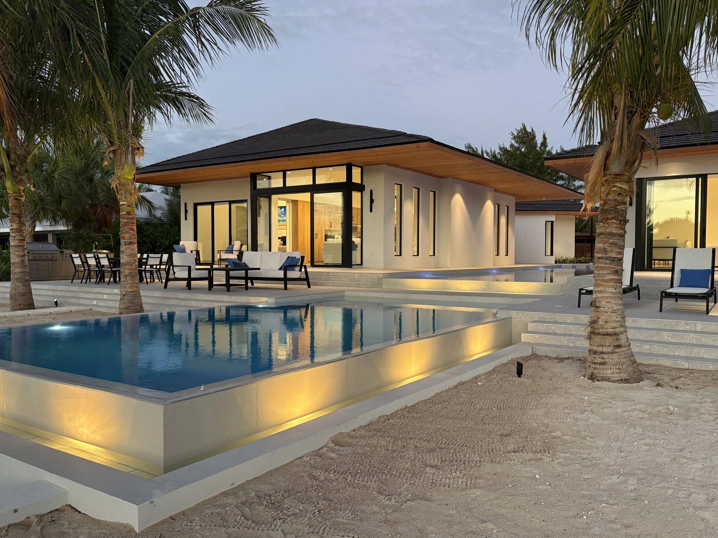 Modern house with large glass windows and doors, outdoor seating area, swimming pool, and palm trees in a tropical setting during dusk.