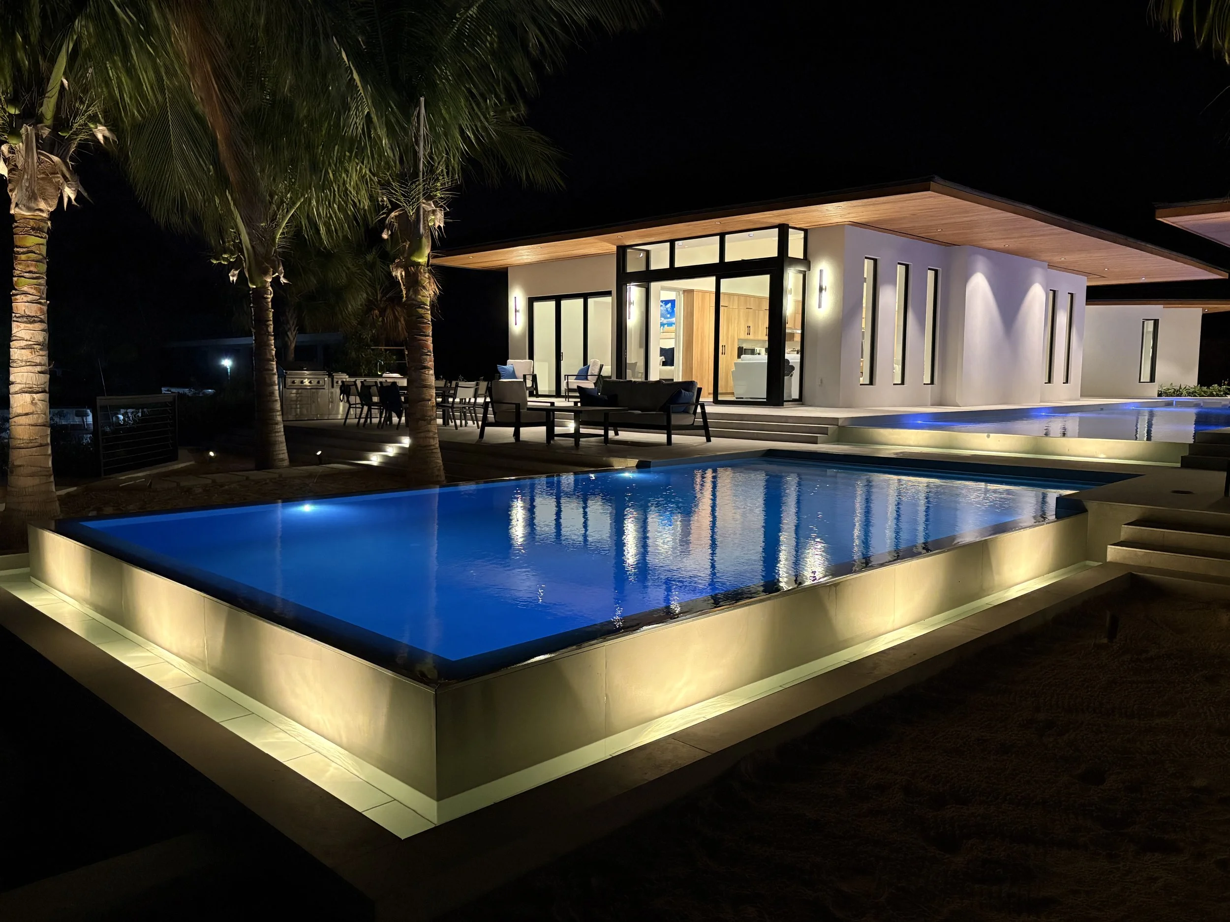 Night view of a modern house with a lit pool in front, surrounded by palm trees and outdoor seating, showcasing contemporary architecture and warm lighting.
