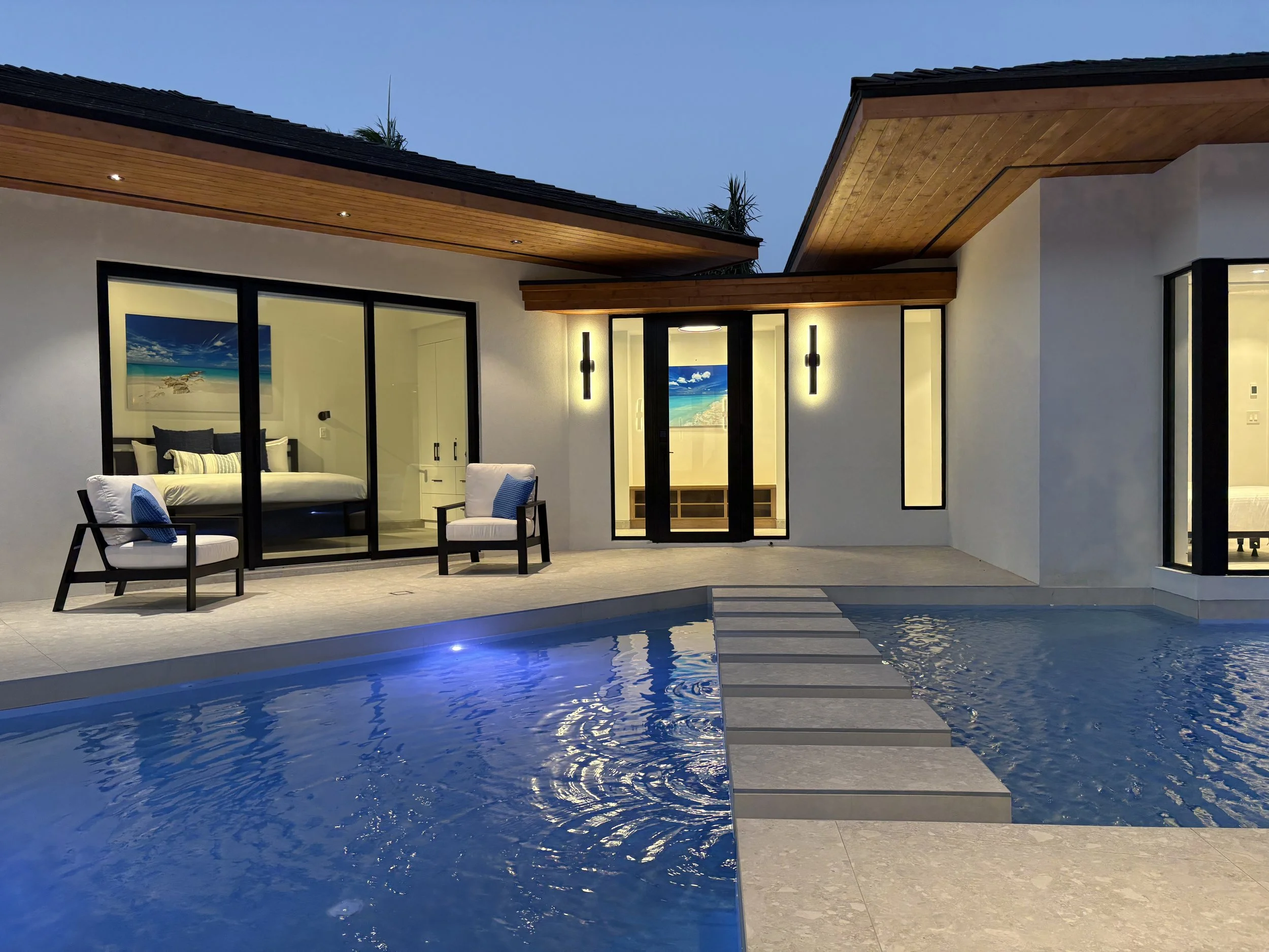 Modern house with a pool outside, illuminated at dusk, with patio furniture and large windows revealing a bedroom inside with a painting of a beach scene.