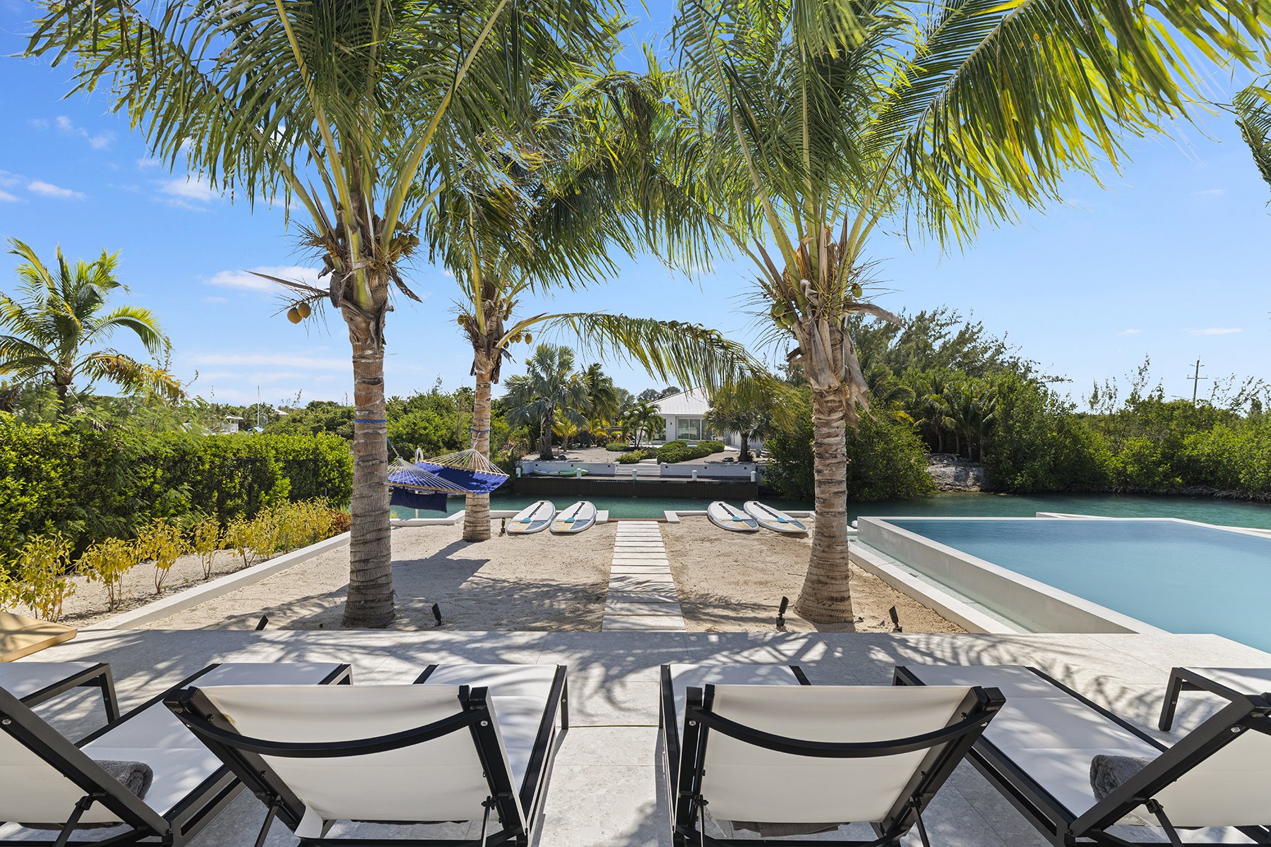 View from a patio with lounge chairs overlooking a tropical beach with palm trees, umbrellas, boats, and a house in the background.