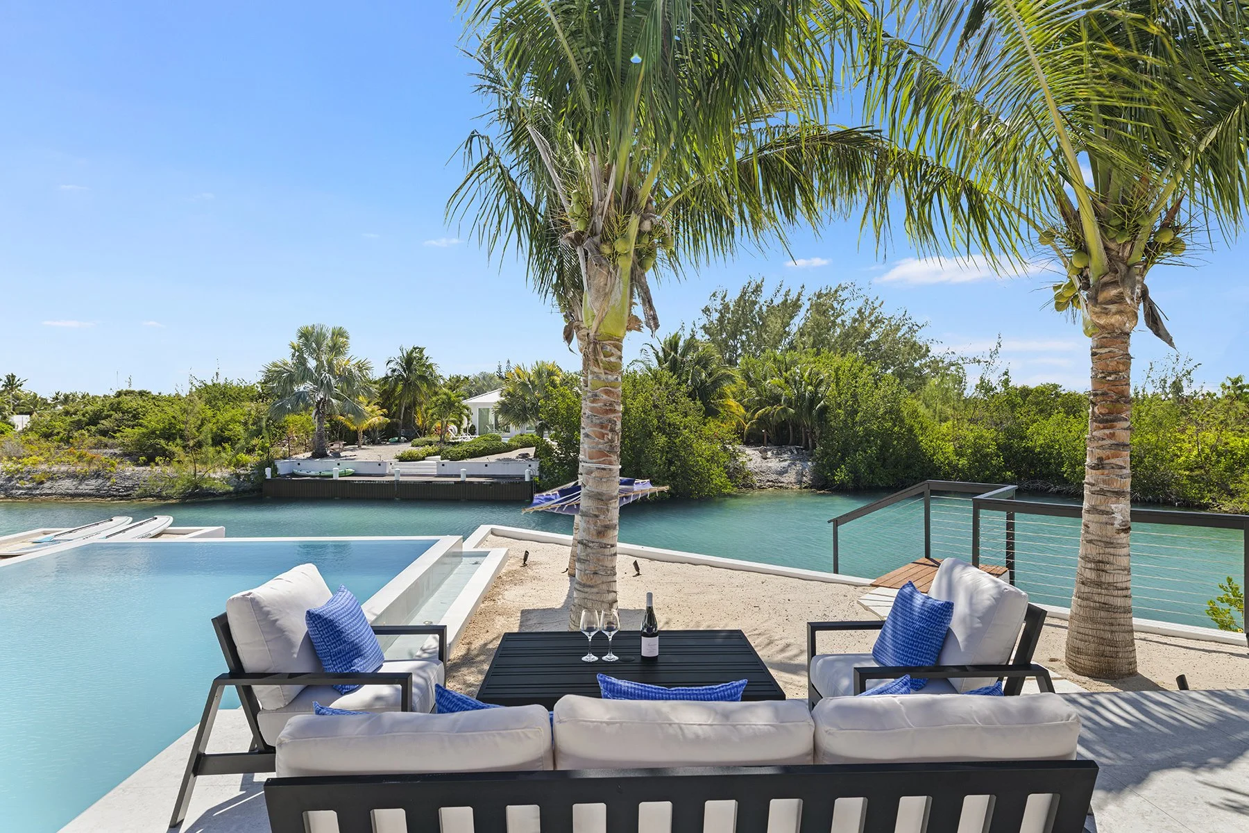 Outdoor patio with white cushioned seating and blue pillows, a black coffee table with a bottle of wine and two glasses, surrounded by palm trees overlooking a body of water and lush greenery under a clear blue sky.