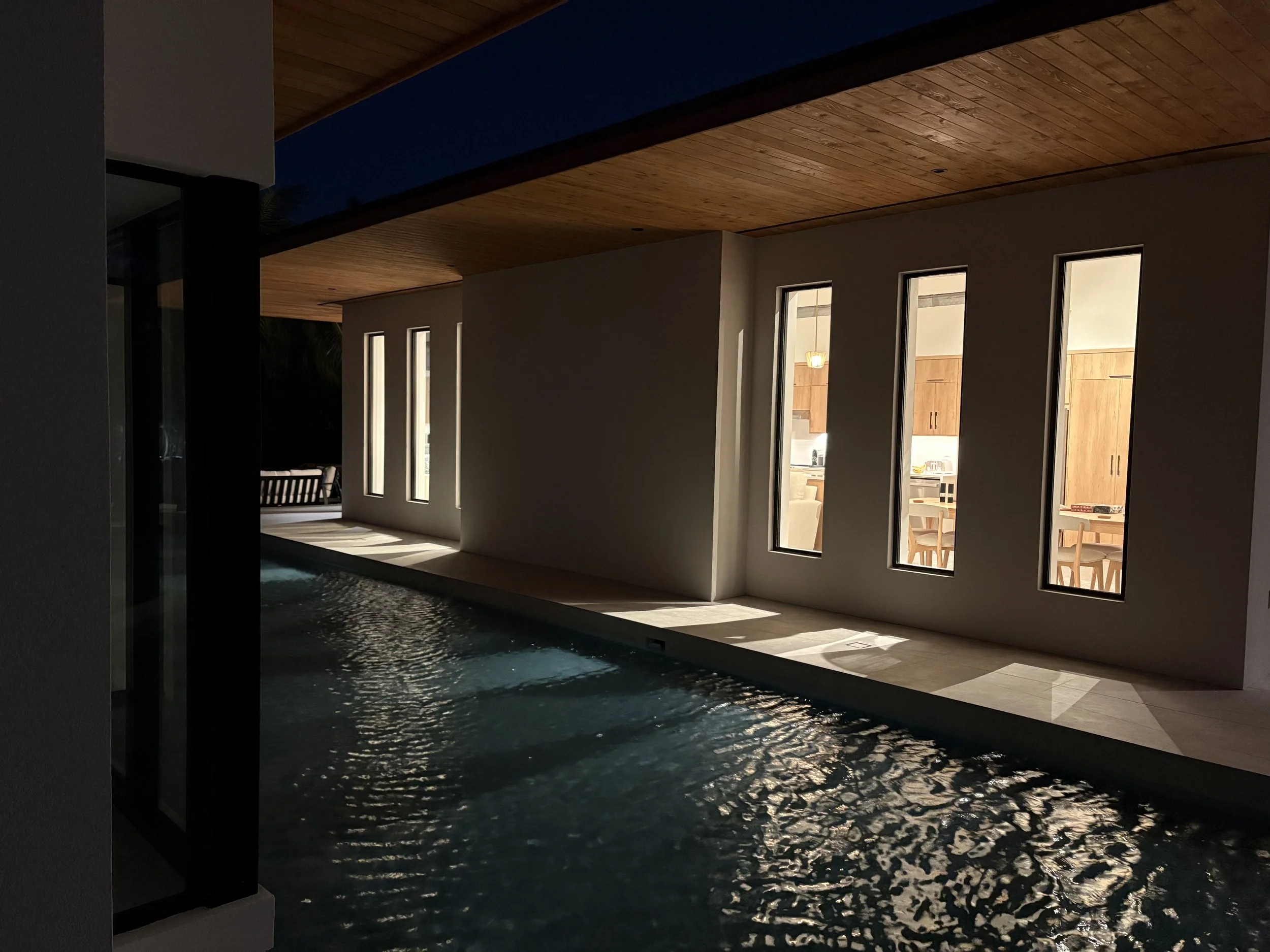 Indoor swimming pool area illuminated at night with a view into a modern kitchen through tall narrow windows.