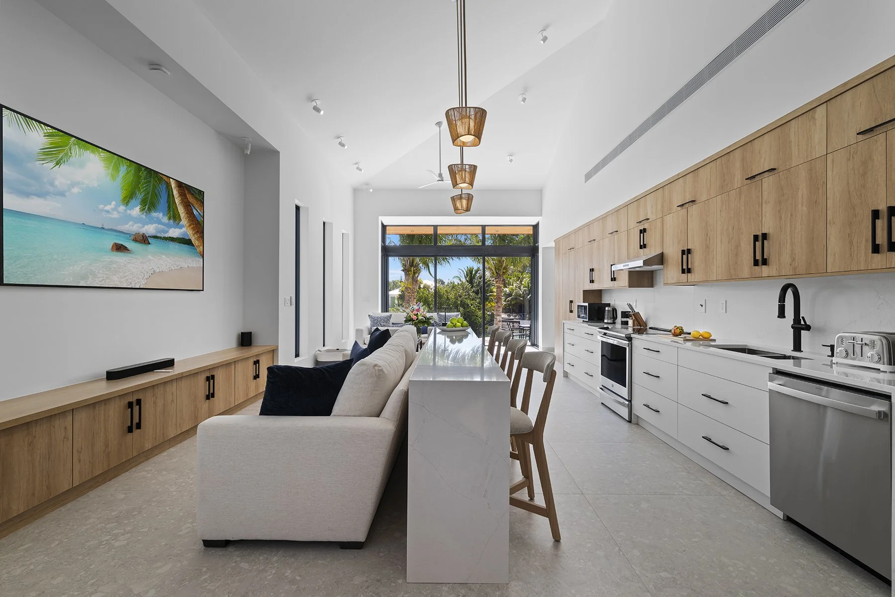 Modern open-concept kitchen and living room with a large window showing palm trees and outdoor scenery, featuring white and natural wood cabinetry, a marble island, and a wall-mounted TV