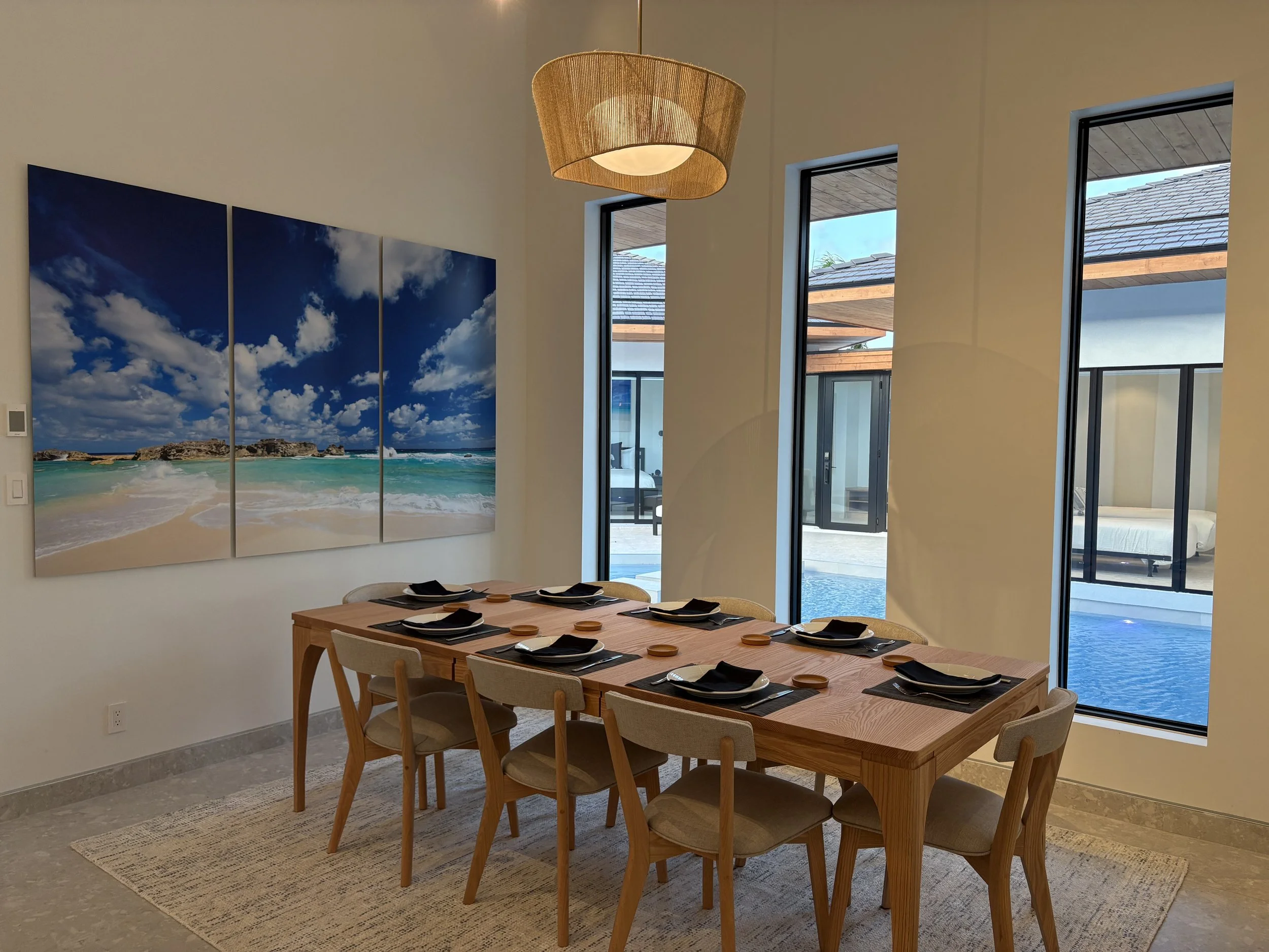 Dining room with a wooden table set for eight, black napkins and plates, and large windows overlooking a pool and patio area.