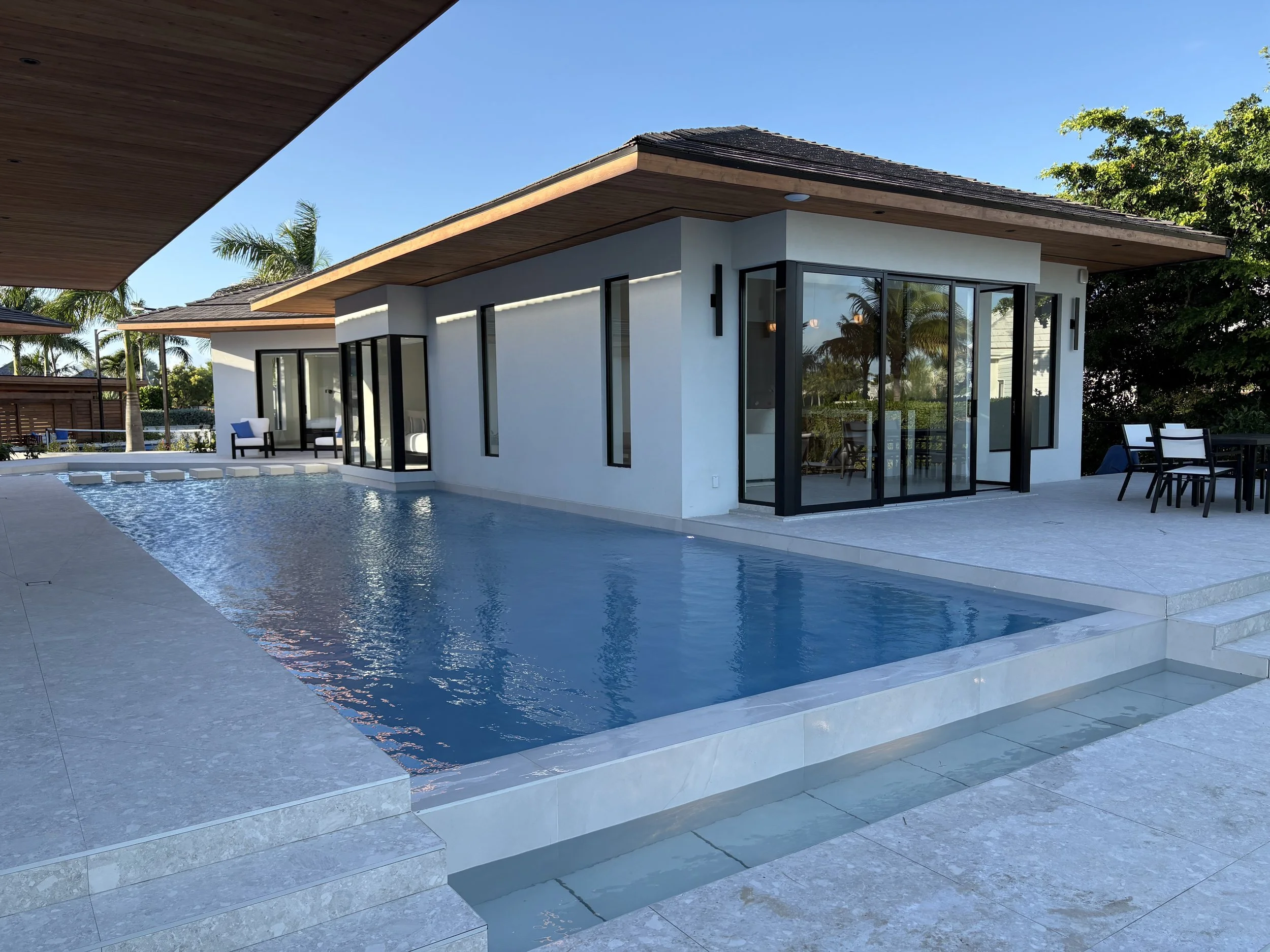 Modern house with a private pool, glass sliding doors, and outdoor seating area, surrounded by tropical trees under a clear blue sky.