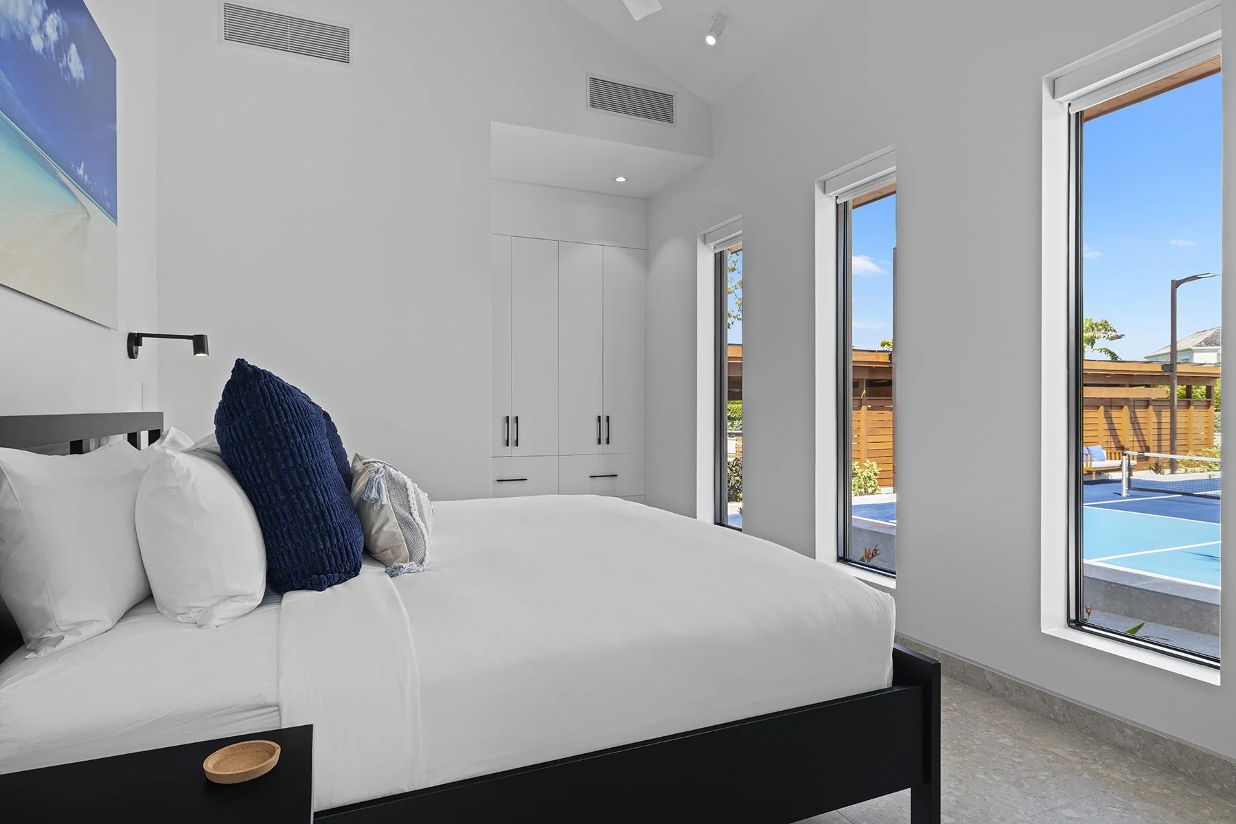 A bedroom with a white bed, blue pillow, and white pillows, three tall windows showing a view of a backyard with a patio and a basketball court, and a built-in white cabinet.