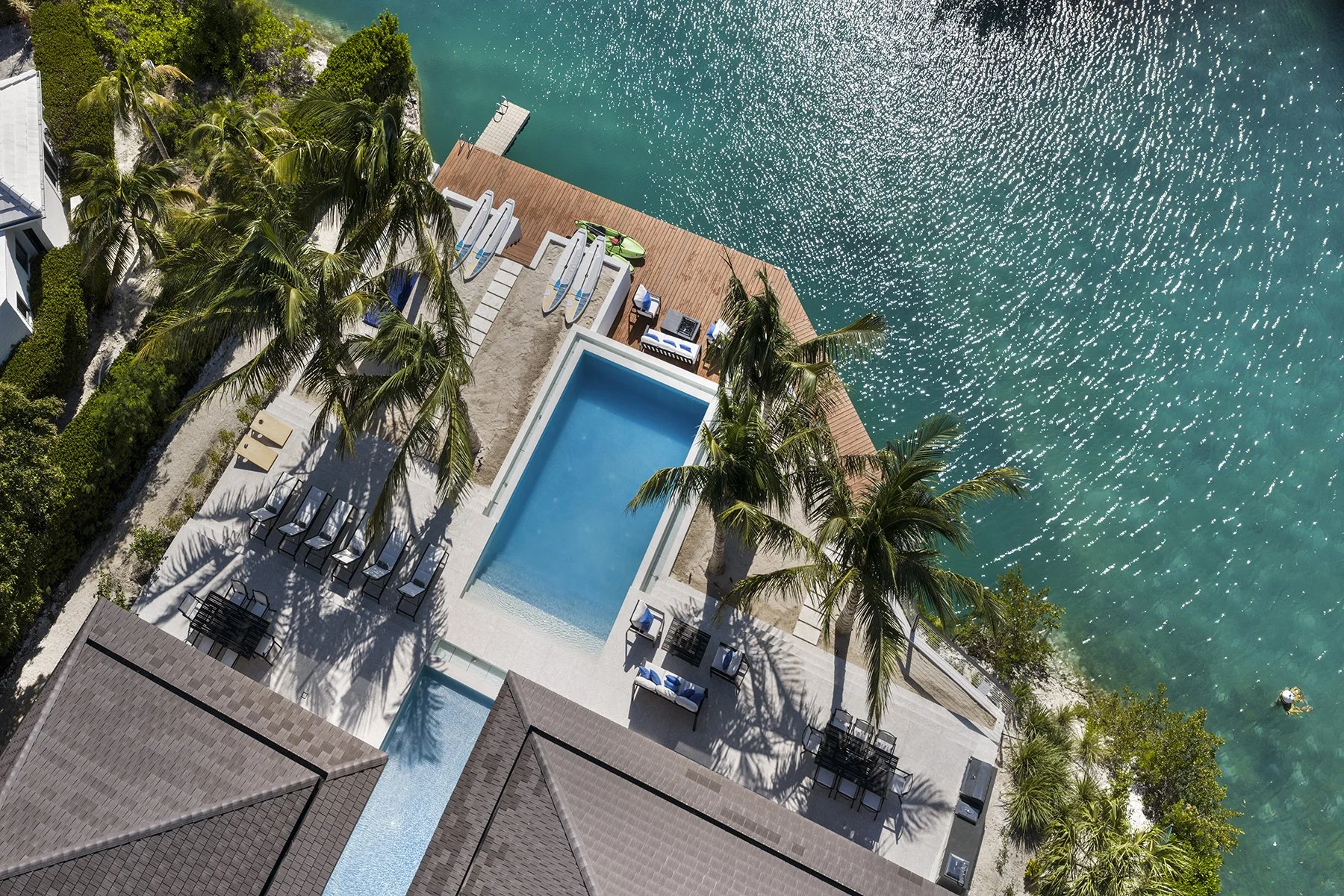 Aerial view of a pool area by the water with palm trees, outdoor seating, and a dock extending into the water.