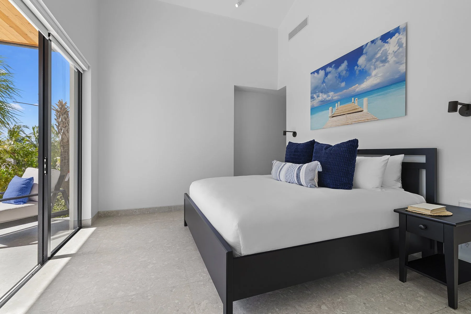Cozy bedroom with a large bed, navy blue and white pillows, a black nightstand with a book, and a large ocean-themed picture on the wall, with sliding glass doors leading to a patio outside.