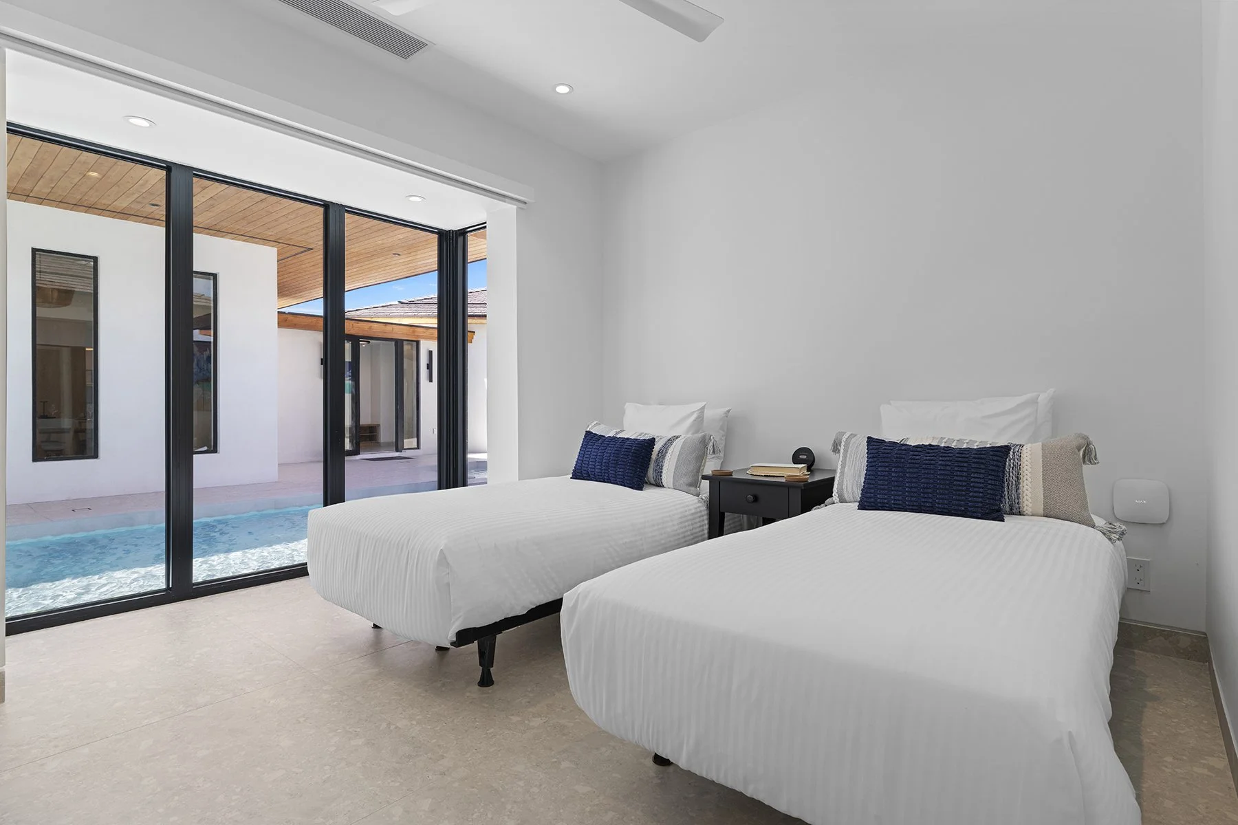 A modern bedroom with two white beds, blue pillows, a nightstand with books and a phone, and a sliding glass door opening to a pool outside.