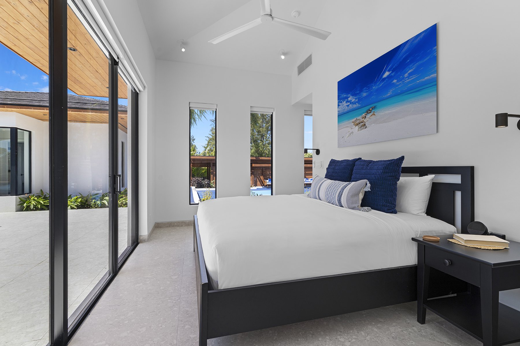 A modern bedroom with white walls, a black bed frame, and a large painting of a beach scene. There is a black bedside table with books and a black lamp. Large glass sliding doors lead to an outdoor patio.