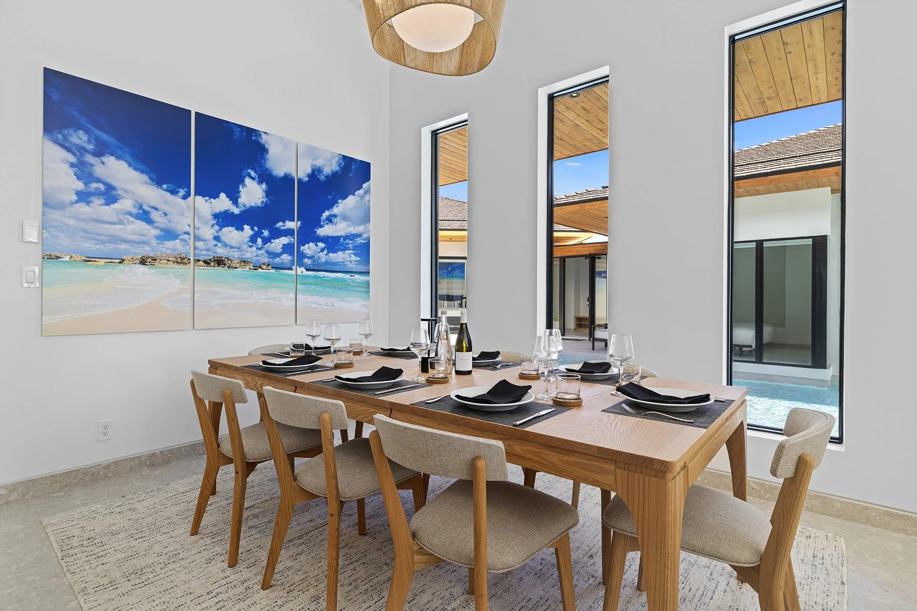 Modern dining room with large windows, a wooden table set for a meal, and beach-themed artwork on the white wall.