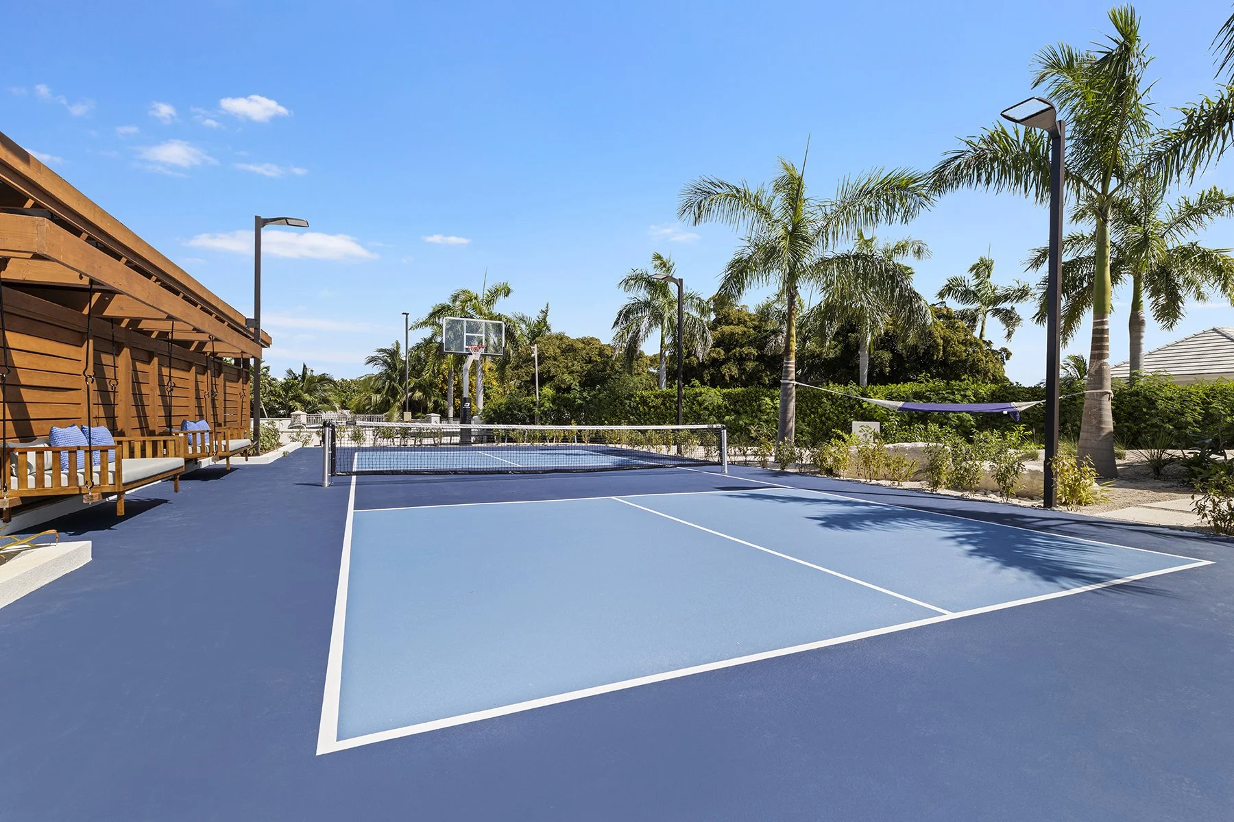 Outdoor tennis court with a volleyball net, surrounded by palm trees and outdoor seating, under a blue sky.
