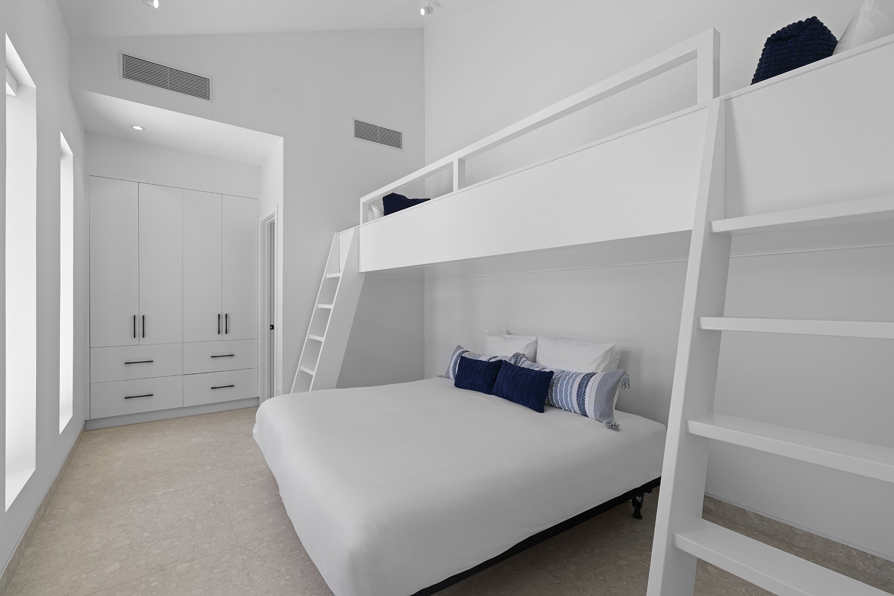 A minimalist bedroom with white walls, a white bed with blue and striped pillows, a built-in closet with black handles, and a ladder leading to a loft area with black pillows.