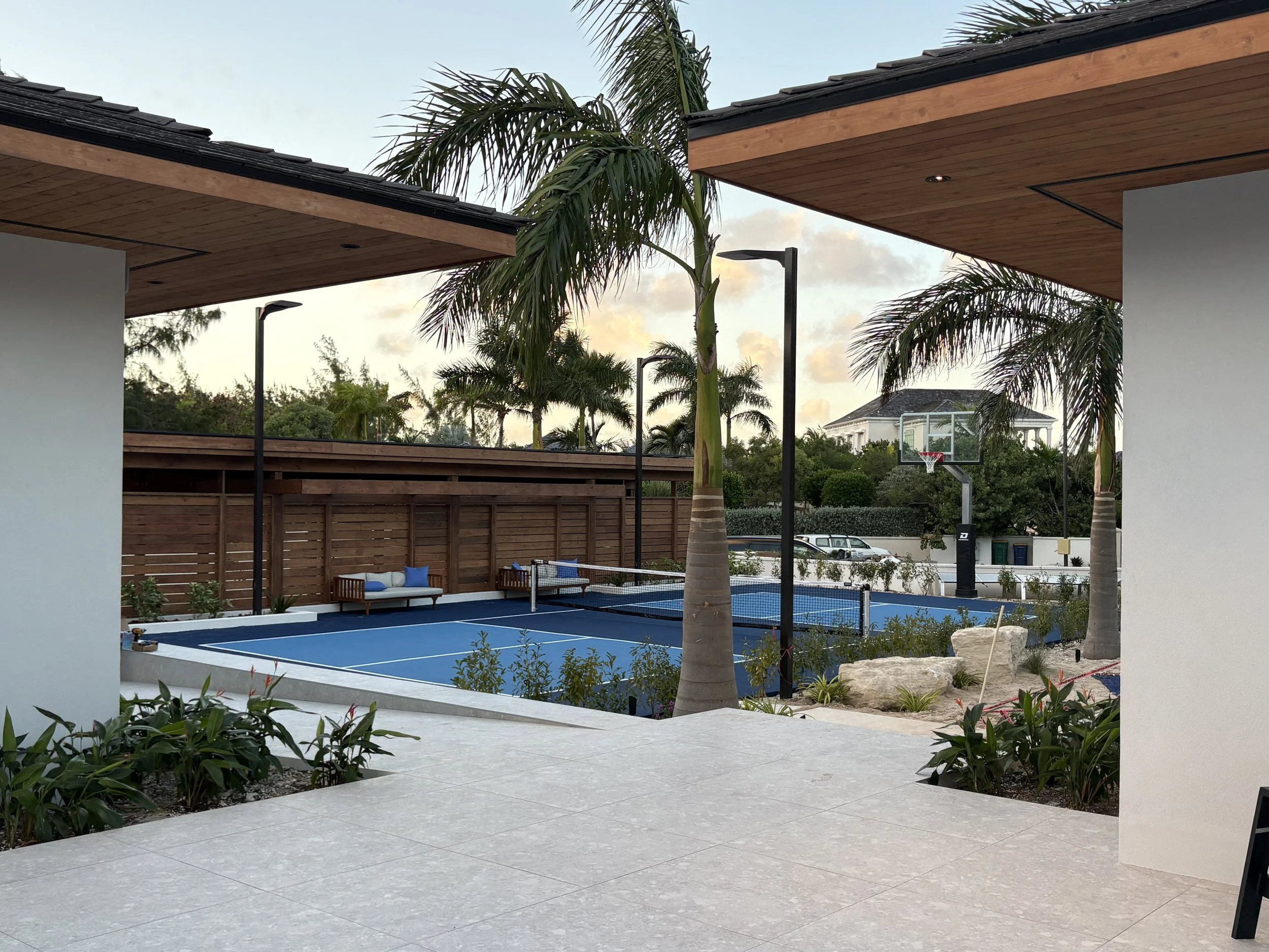 Modern outdoor patio overlooking a backyard basketball court with palm trees and benches, surrounded by a wooden fence and lush greenery.