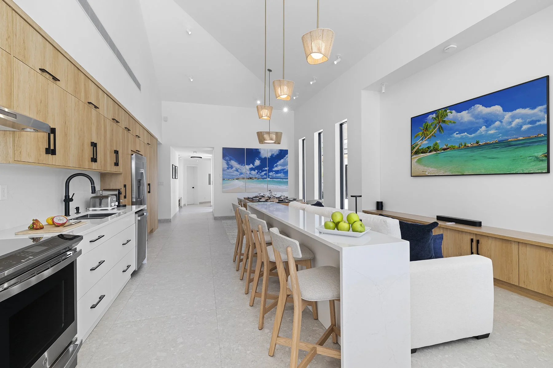 Open-concept living and dining area with white walls, large windows, wooden and white cabinetry, a kitchen island with bar stools, a white sofa with blue pillows, wall-mounted flat-screen TVs displaying beach scenes, and modern lighting fixtures.