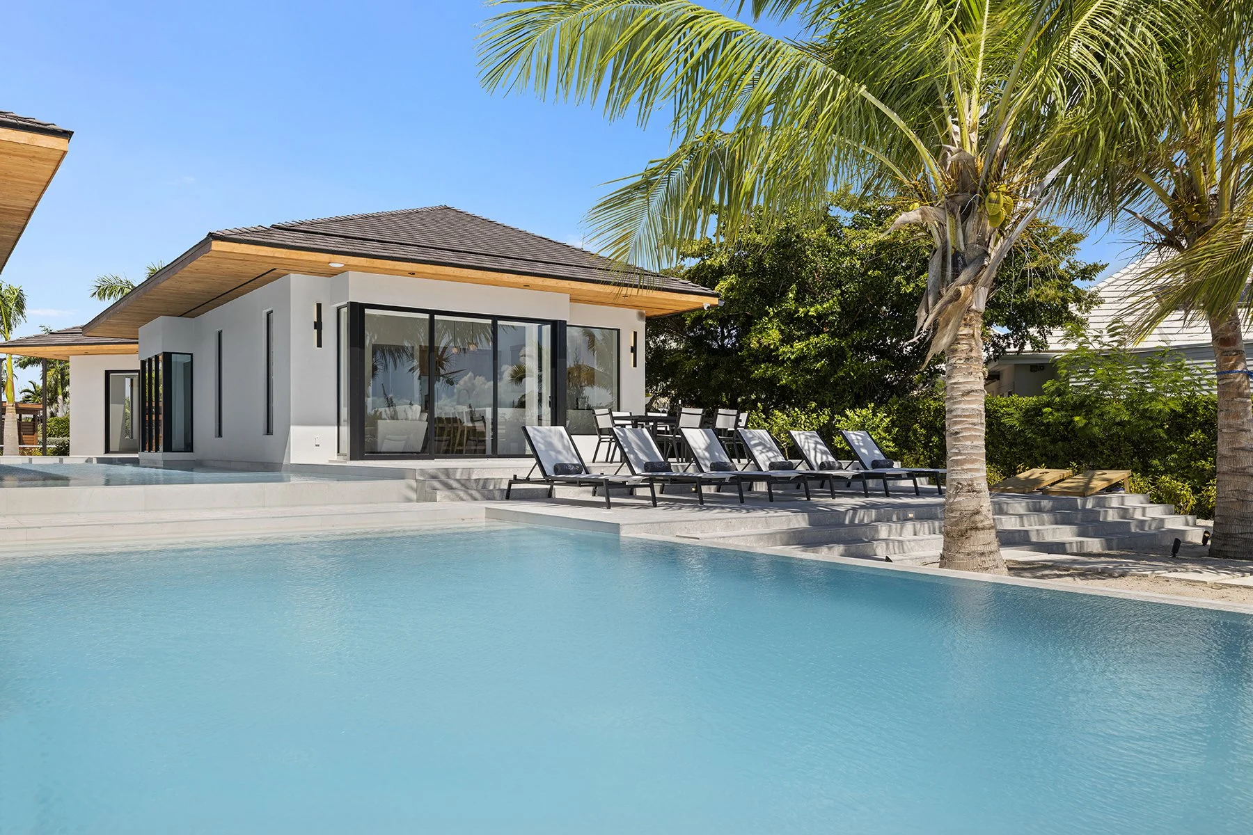 Luxury modern house with large sliding glass doors, outdoor pool, palm trees, and lounge chairs under a blue sky.