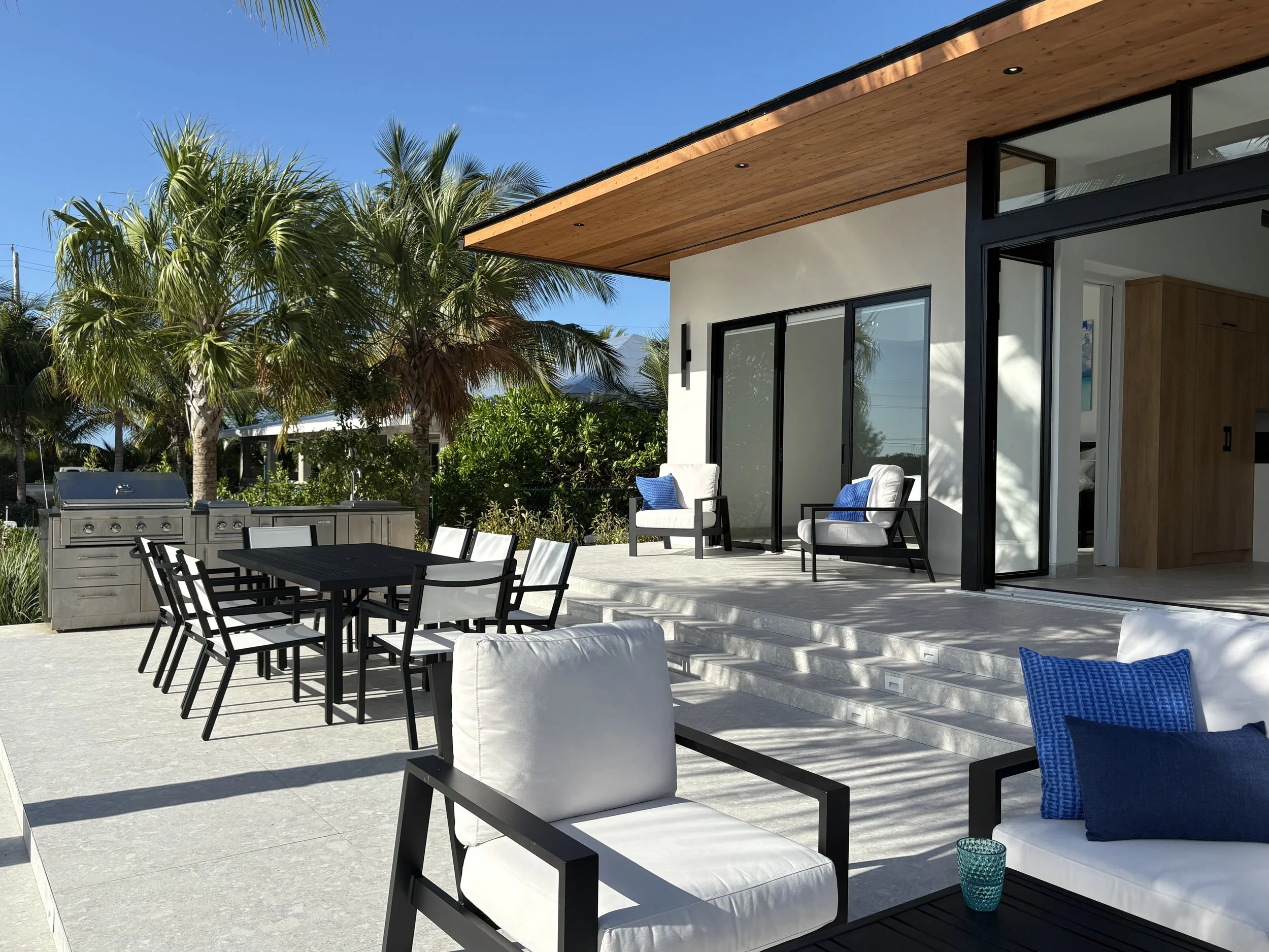 Modern outdoor patio with white lounge chairs, a dining table with chairs, and a built-in grill, surrounded by palm trees and lush greenery.