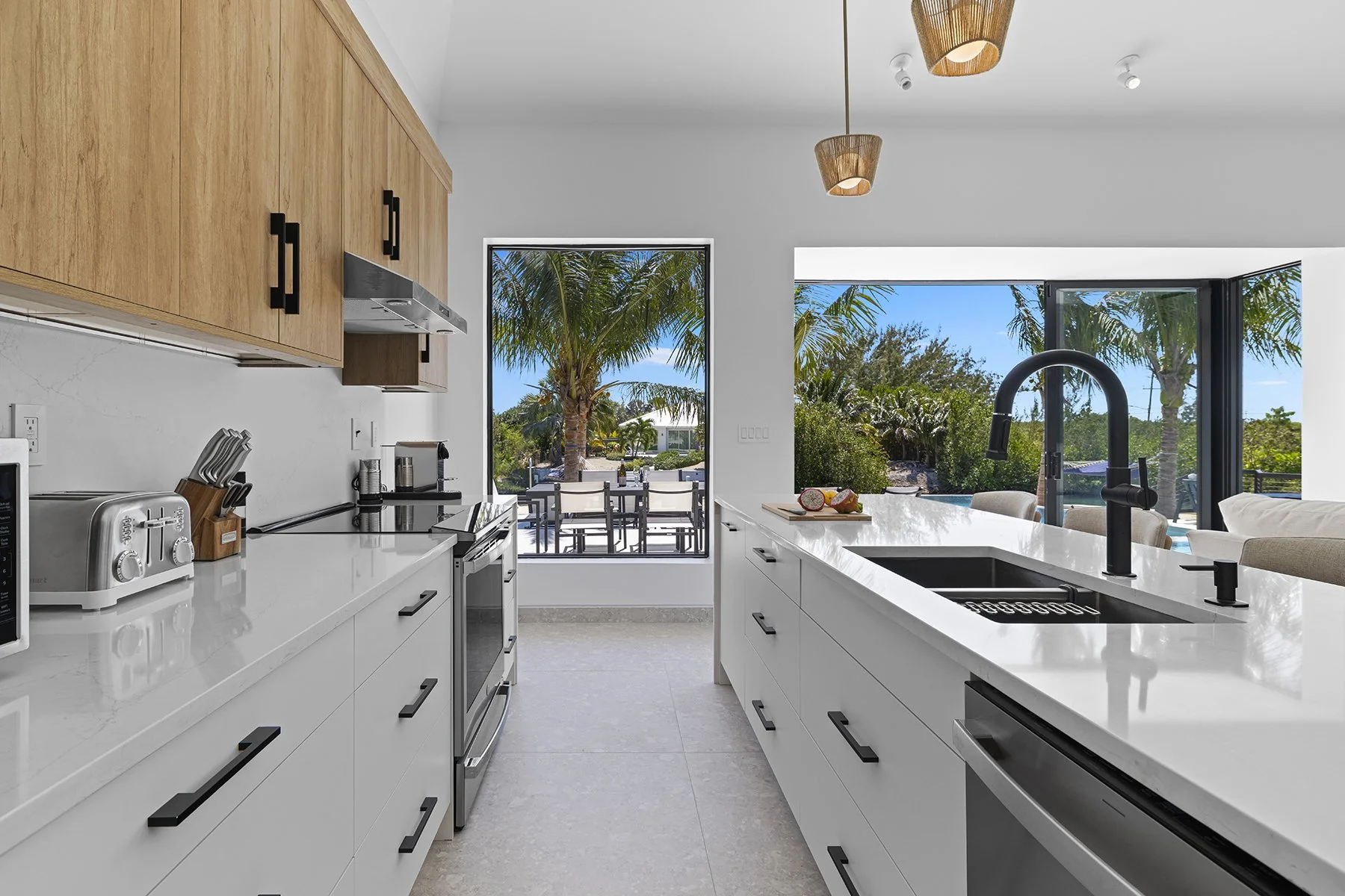 Modern kitchen with white countertops, black sink faucet, wooden cabinets, and large windows overlooking palm trees and outdoor patio area.