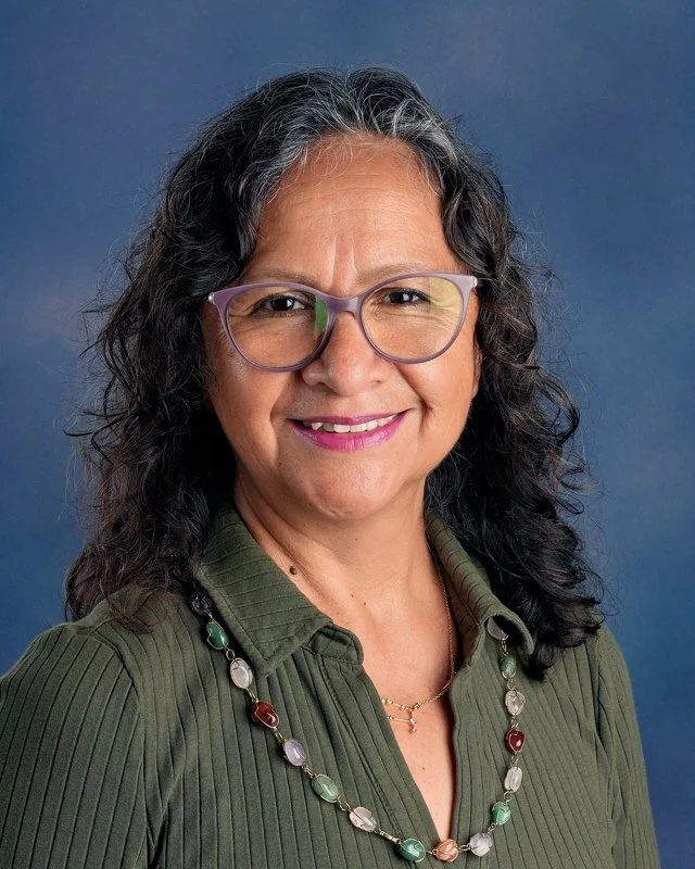 A woman with curly black hair, glasses, and a gentle smile, wearing a green blouse with ruffled details, standing against a blue background.