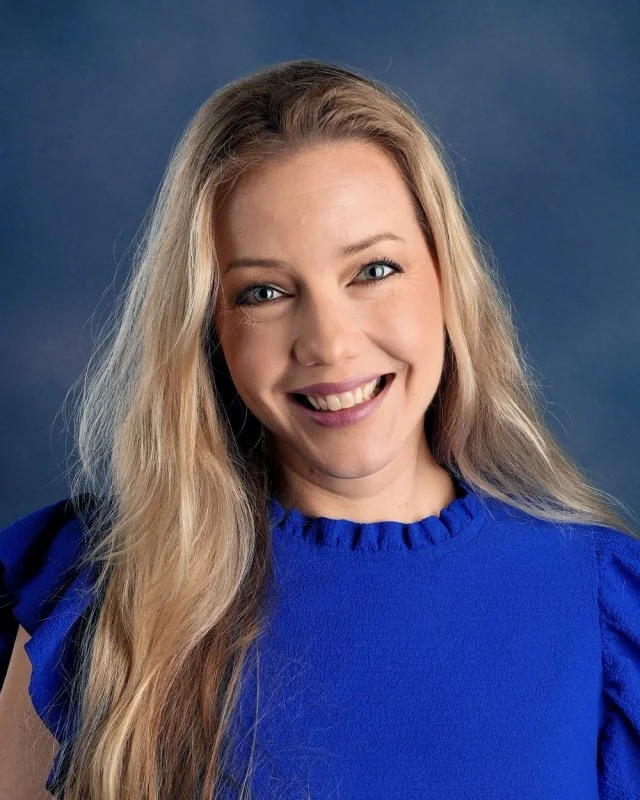 A woman with blue eyes, and fair skin smiling, wearing a black polo shirt with a logo and text on it, against a blue background.