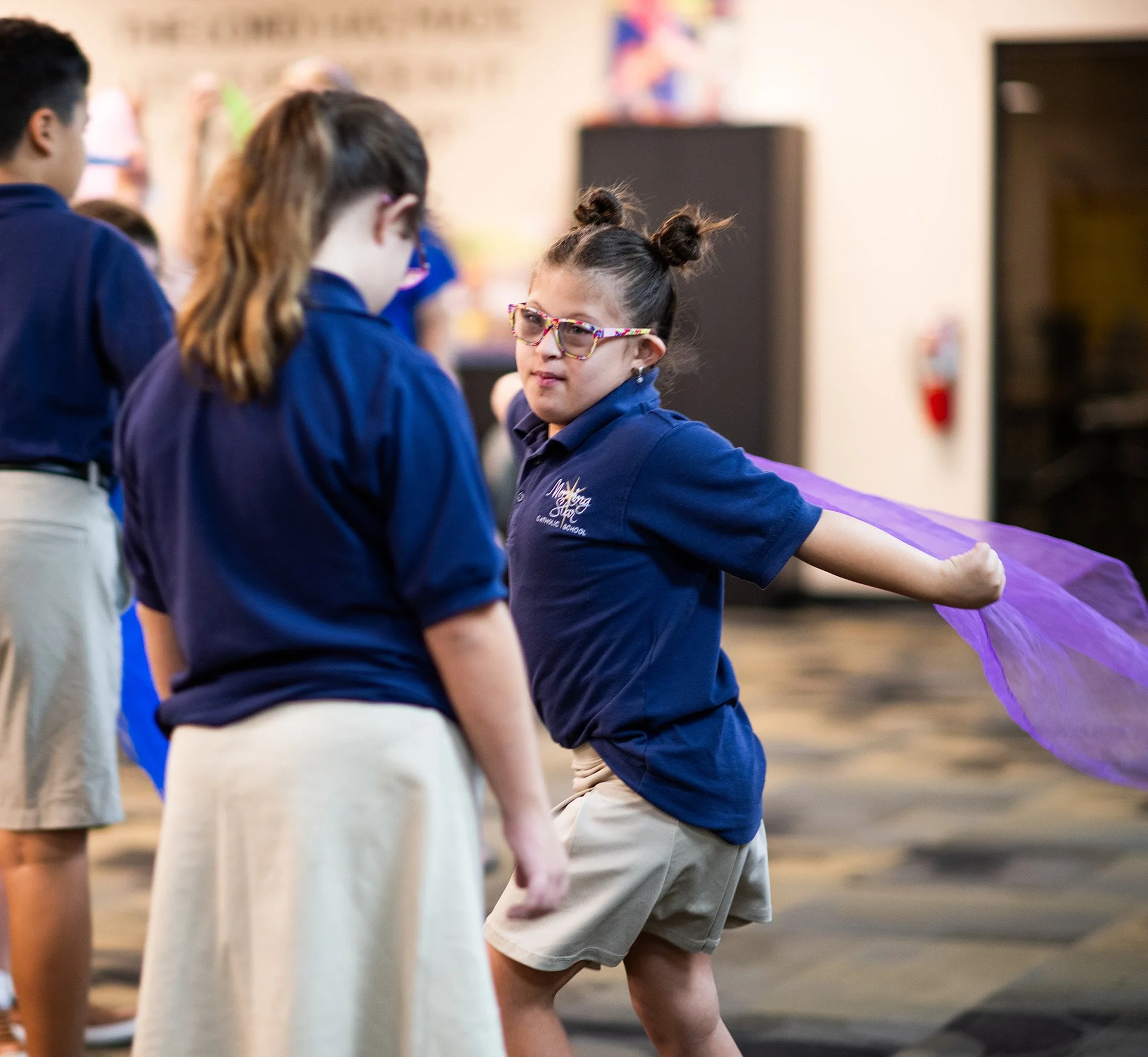 A young girl wearing glasses and a navy blue school uniform is whirling a purple scarf in the air, with other students around her in a classroom or school hall.