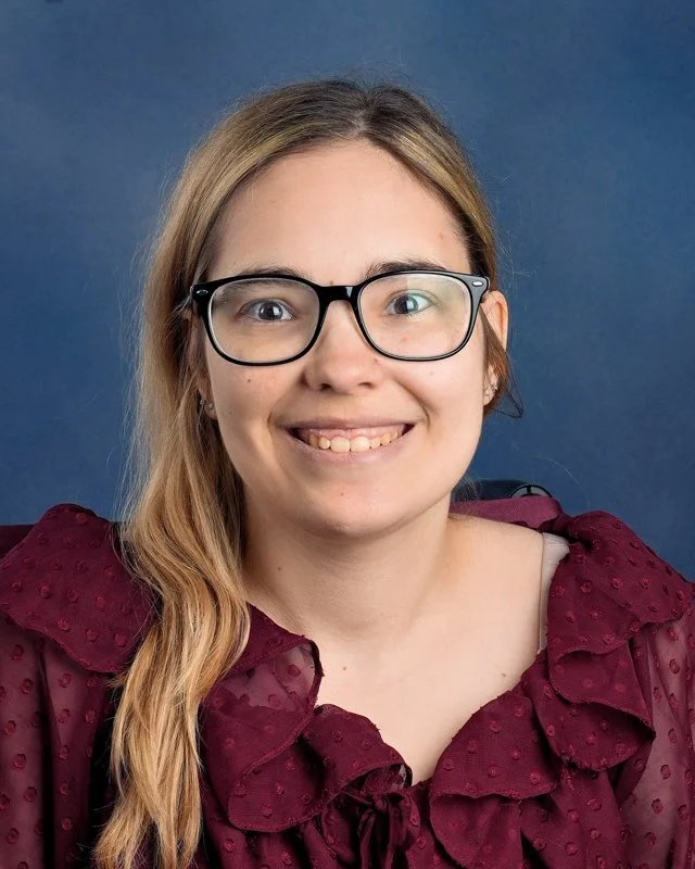 Portrait of a young woman with long, light brown hair, wearing glasses and a navy blue floral blouse, smiling against a blue background.
