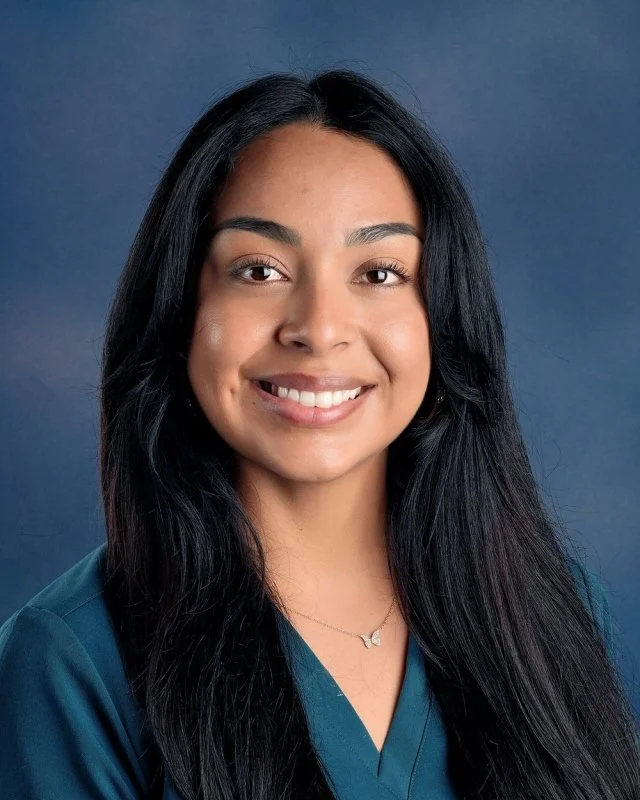 A woman smiling at the camera with shoulder-length gray and light brown hair, wearing a navy blue collared shirt with a necklace, against a blue background.
