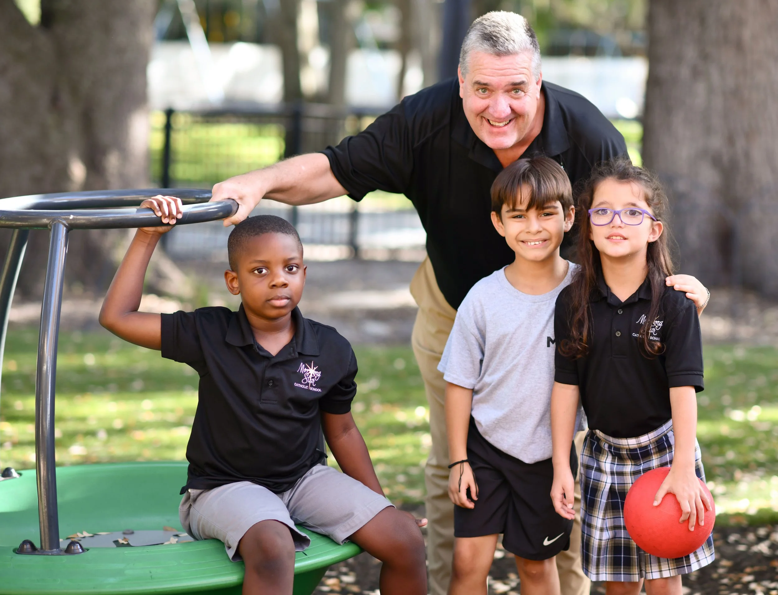 A group of boys in a line with an older man, all in various stages of bouncing a basketball. The court is in front of a playground, and it is a bright, sunny day.