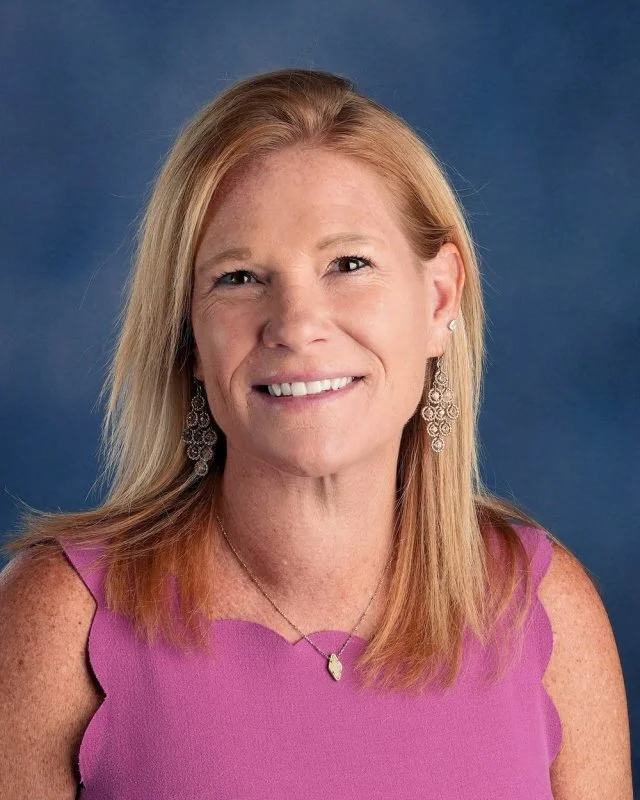A woman with blonde hair and light skin, wearing teal earrings and a dark blue top, smiling against a blue background.