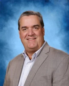 Professional headshot of a smiling man in a light grey suit and white shirt against a blue background.