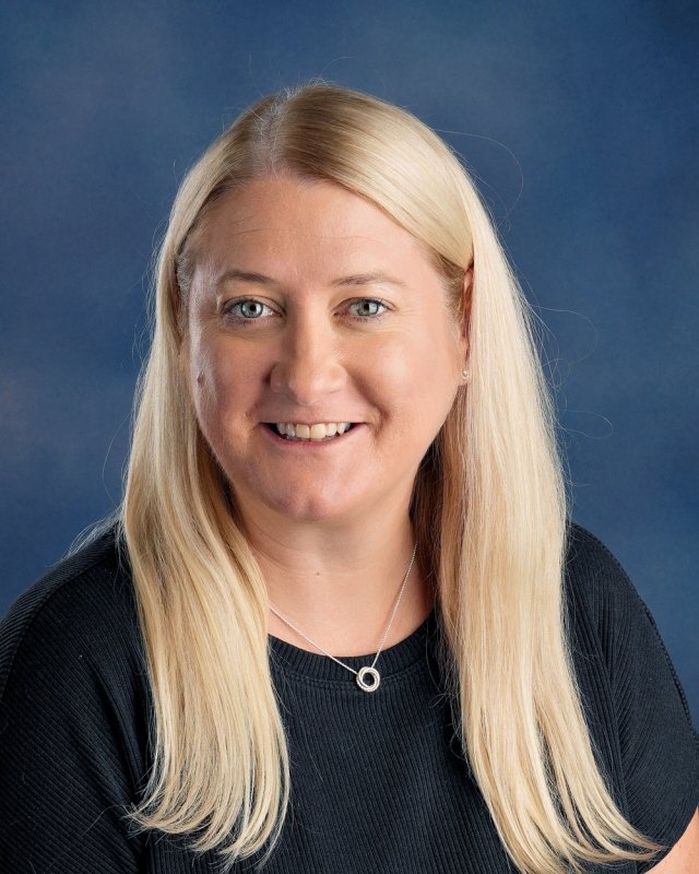 A woman with long blonde hair and a black top smiling in front of a blue background.